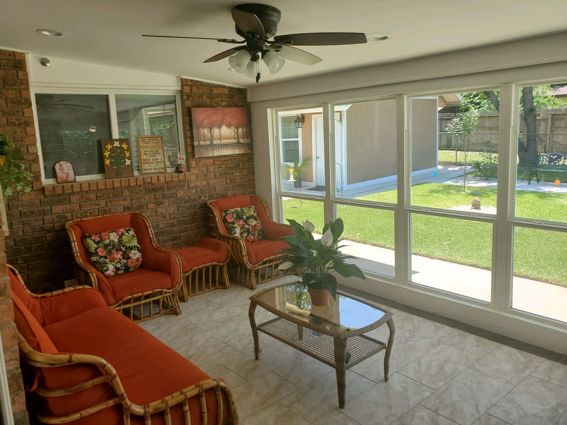 A bright sunroom with large windows overlooking a green lawn and garden. The room features a ceiling fan, brick wall, and wicker furniture with red cushions and floral pillows. A glass-top wicker coffee table holds a potted plant.