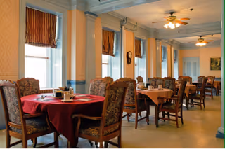 A dining room with several wooden tables covered with tablecloths, surrounded by upholstered chairs. The room has large windows with brown curtains, beige patterned wallpaper, and ceiling fans with lights. The tables are set with condiments and napkins.