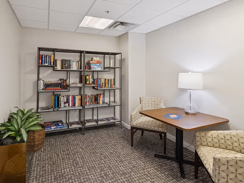 A small reading or sitting area with a metal bookshelf filled with books against a beige wall. There are two patterned armchairs around a wooden table with a lamp on it. A potted plant is placed on the floor near the bookshelf. The room has a carpeted floor and a drop ceiling with fluorescent lighting.