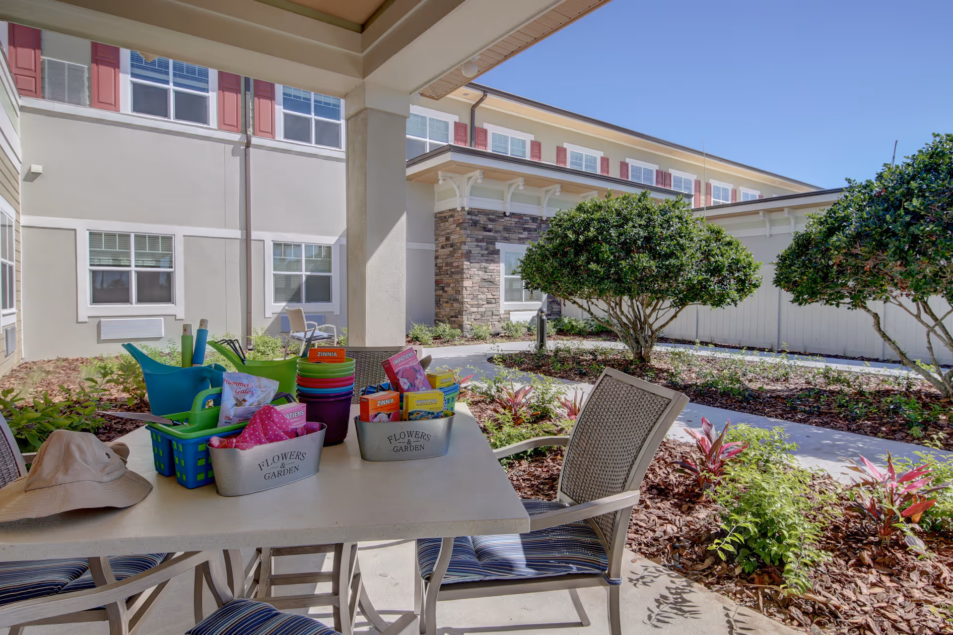 Outdoor covered patio area at Beach House Assisted Living & Memory Care with a table and chairs. On the table are gardening supplies including colorful pots, seed packets, a watering can, and a sun hat. The patio overlooks a landscaped garden with bushes, plants, and a walking path. The building exterior with windows and red shutters is visible in the background under a clear blue sky.