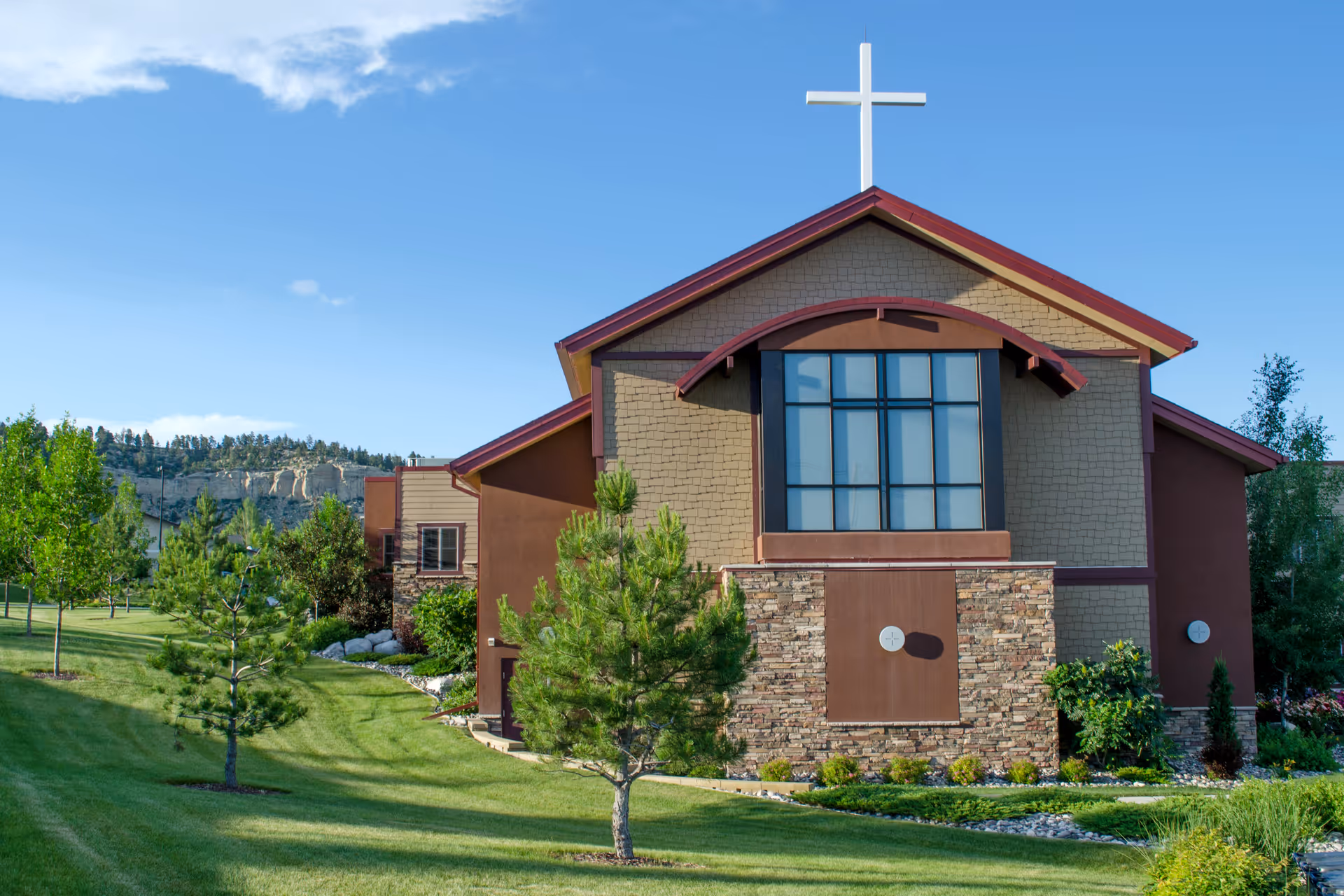 Exterior view of a building with a large window and a white cross on the roof, surrounded by green grass, trees, and shrubs under a clear blue sky.