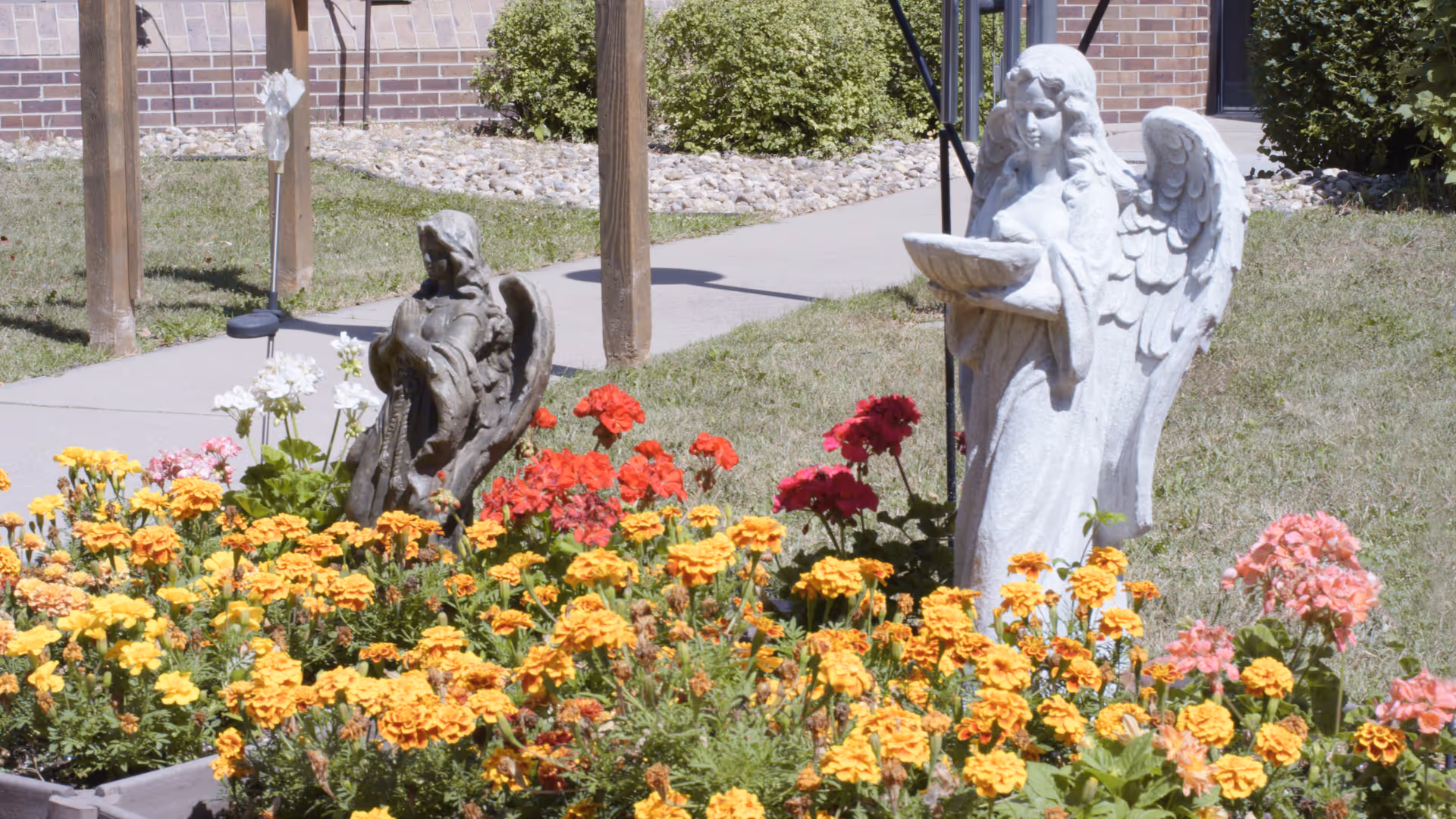 A garden area with colorful flowers including yellow, orange, red, pink, and white blooms. Two angel statues are placed among the flowers, one dark-colored and one white, with a brick building and a sidewalk in the background.