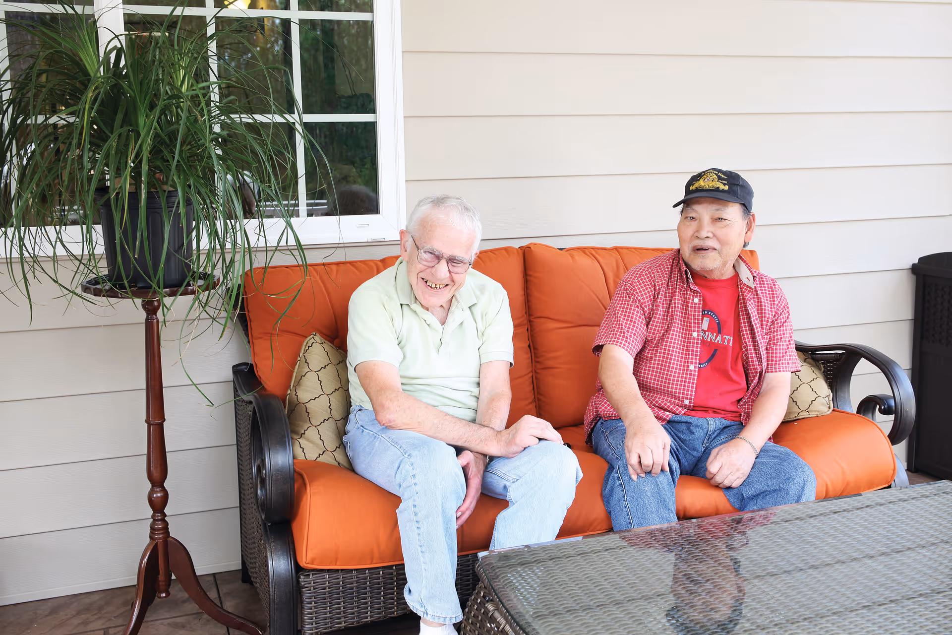Two elderly men sitting on an orange-cushioned outdoor sofa on a patio with a potted plant and glass-top table.