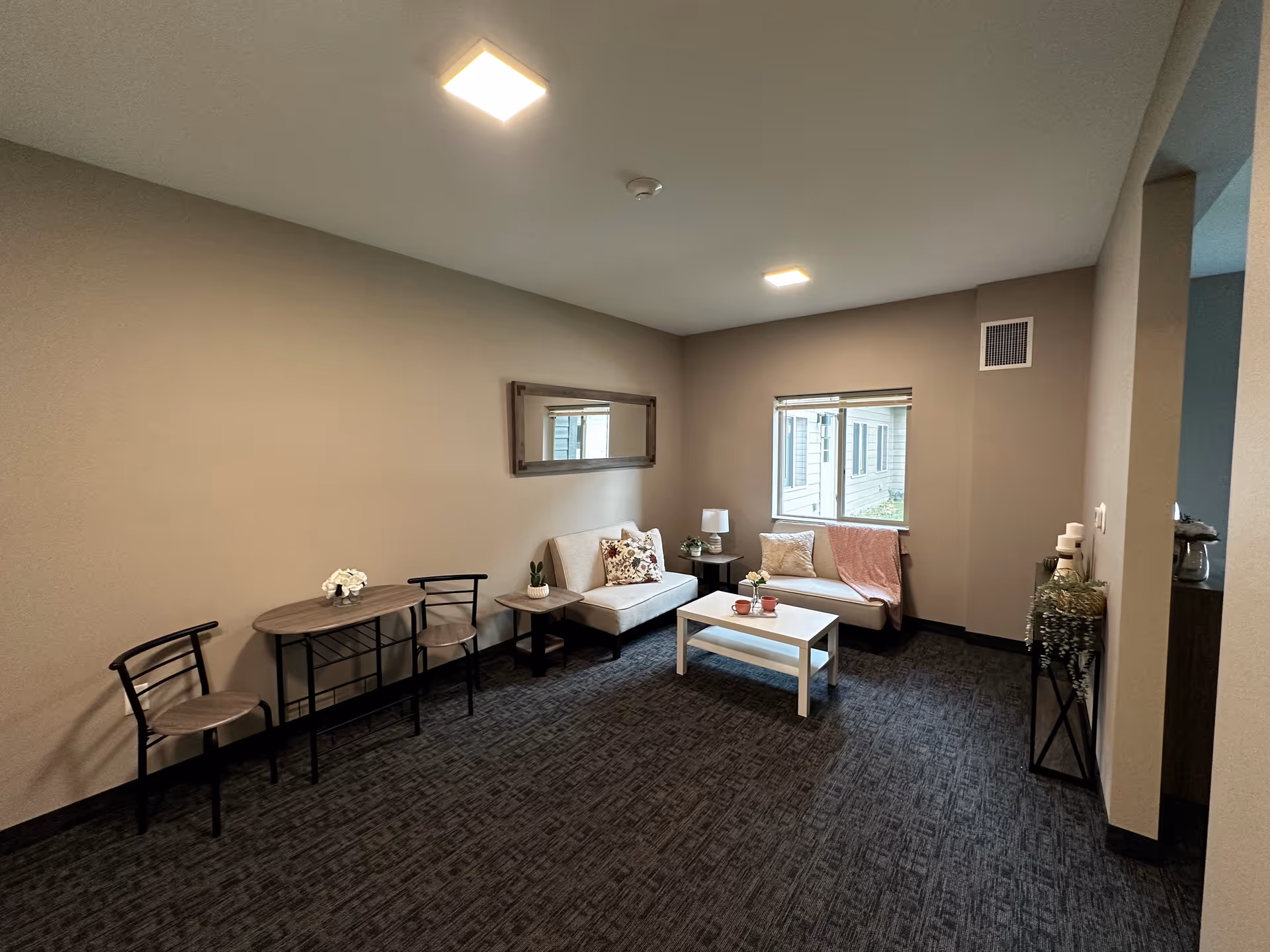 A cozy living room area with two beige sofas, a white coffee table with two cups and a small flower vase, a side table with a lamp, and a small table with two chairs. The room has a window with a view of the outside building, a large mirror on the wall, and carpeted flooring.