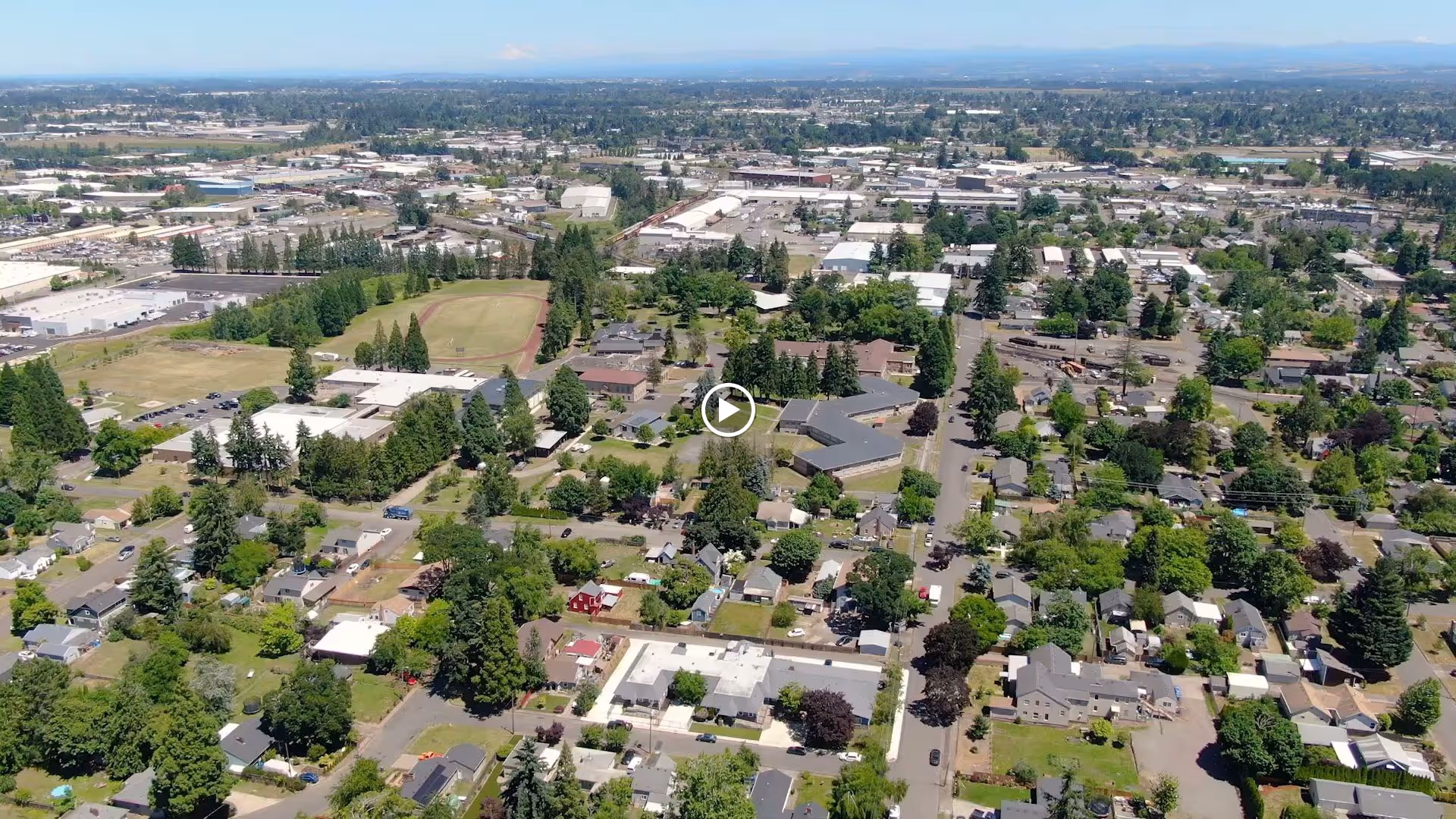 Aerial view of a suburban neighborhood with numerous houses, trees, and streets. In the distance, there are industrial buildings and a sports field with a baseball diamond. The landscape extends to the horizon under a clear blue sky.