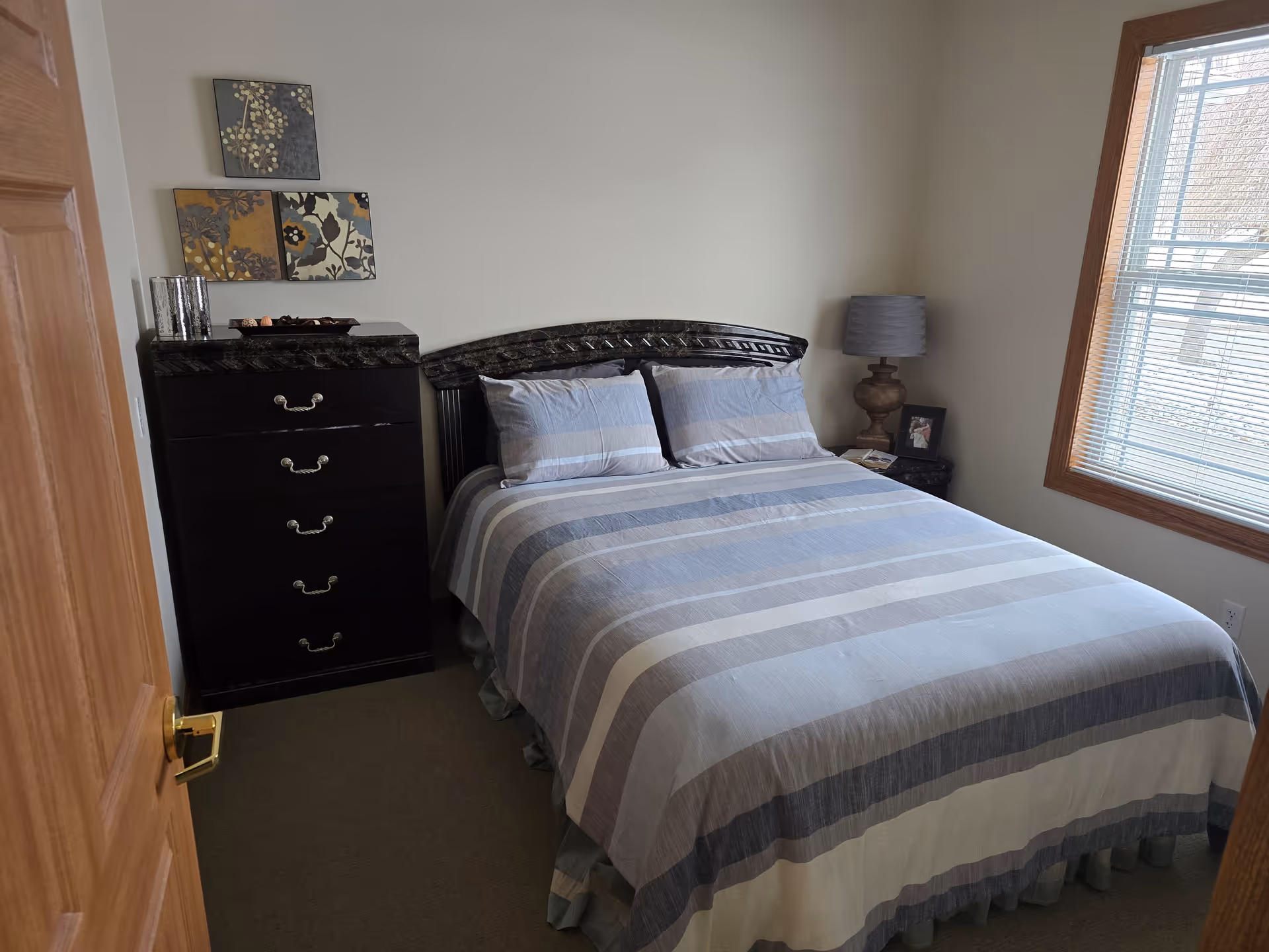 Bedroom with a striped bed, dark wooden headboard and dresser, a lamp on a nightstand, and a window with blinds.