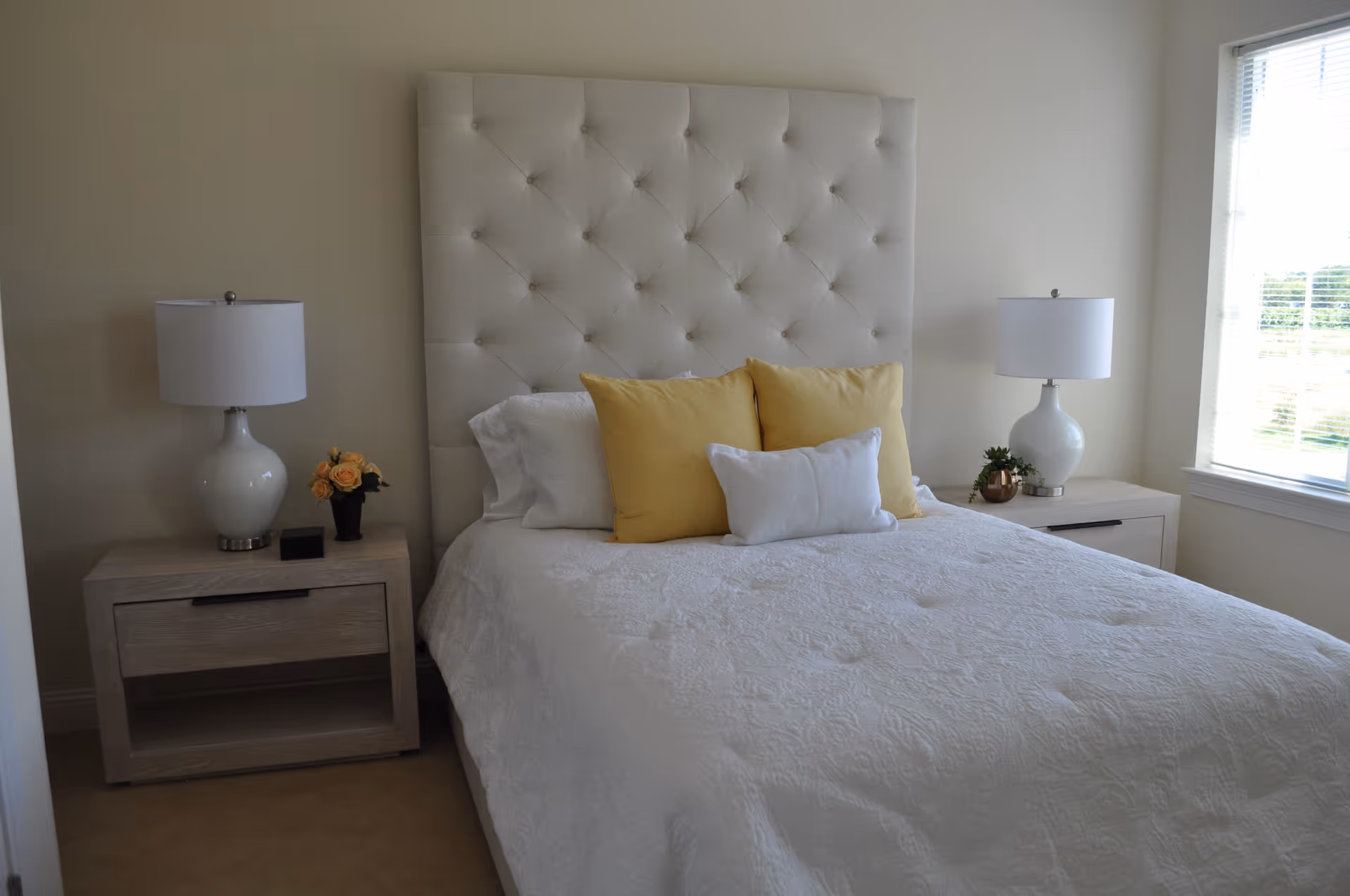 A bedroom with a large white tufted headboard, a bed with white bedding and three pillows (two yellow and one white). There are two matching wooden nightstands on either side of the bed, each with a white lamp. The left nightstand also has a small vase with yellow flowers, and the right nightstand has a small plant. A window with blinds is on the right side of the room, letting in natural light.