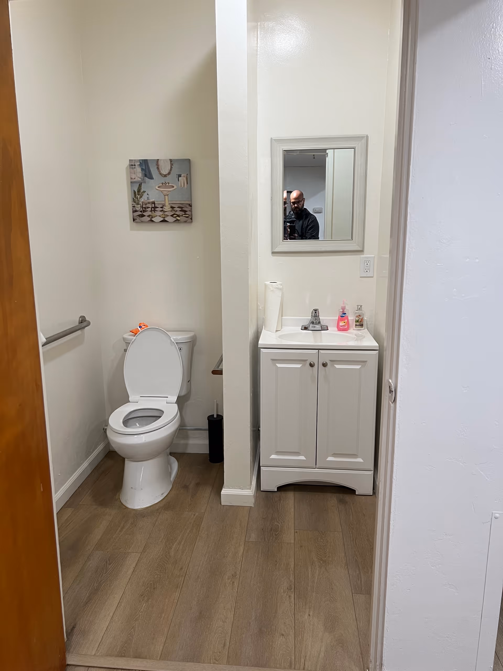 A small bathroom with a white toilet with the lid open, a white vanity with a sink and faucet, a mirror above the sink, a roll of paper towels, hand soap, and a bottle of lotion on the vanity. There is a painting on the wall above the toilet and a grab bar on the left wall. The floor is wood-style laminate.