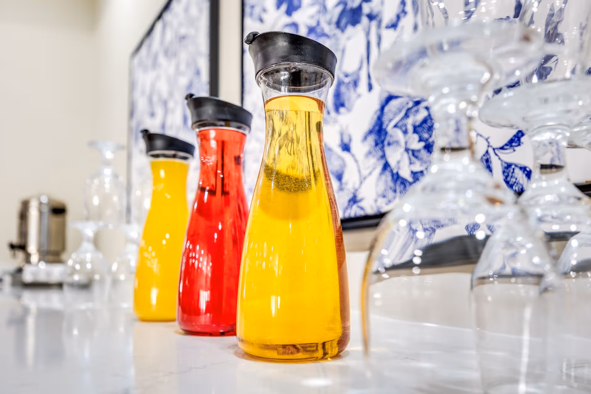 Three glass carafes filled with yellow and red beverages lined up on a white countertop with upside-down wine glasses nearby and a blue and white floral artwork in the background.