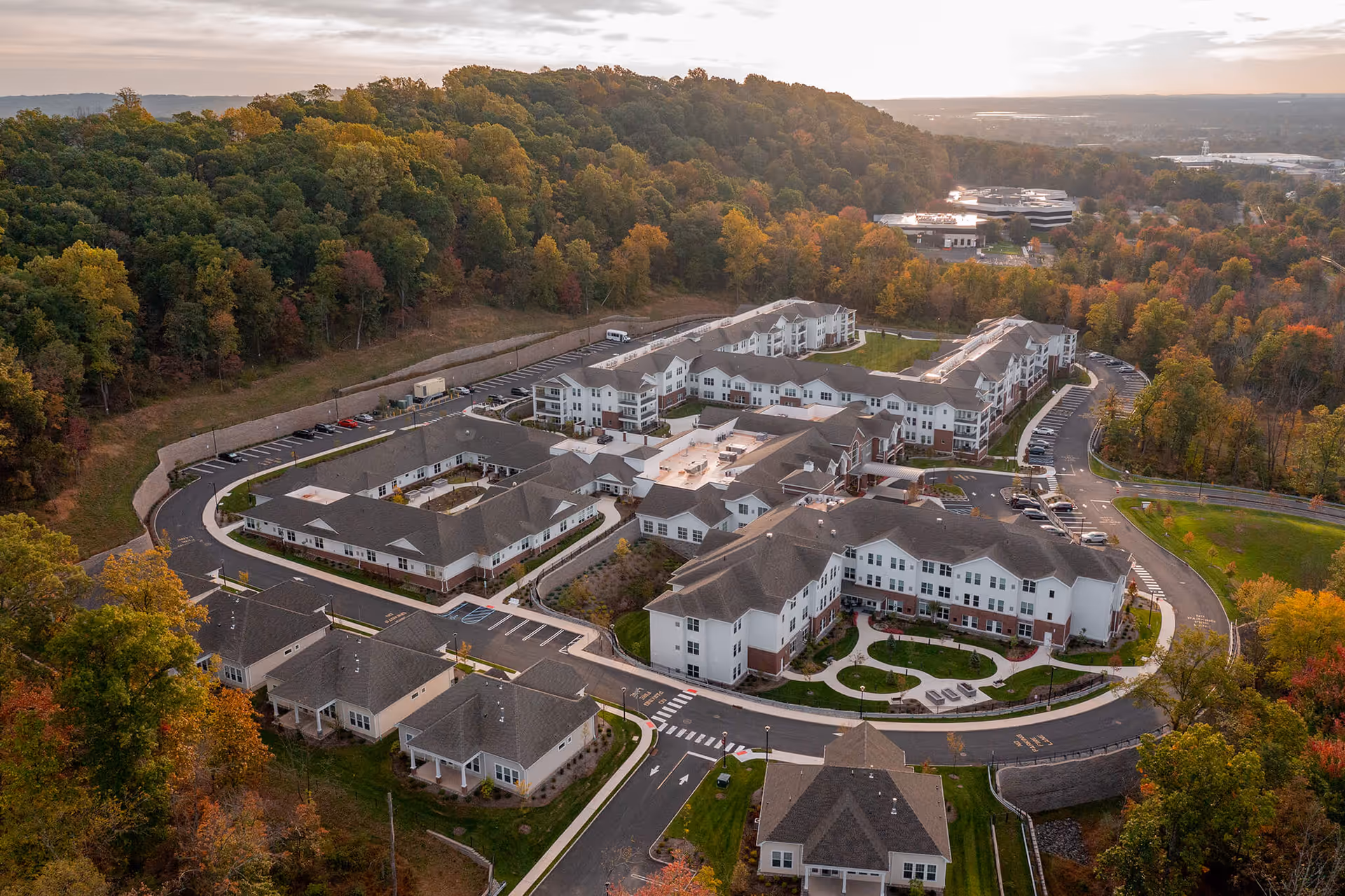 Aerial view of The Delaney of Bridgewater senior living facility surrounded by trees with autumn foliage, showing multiple white buildings with gray roofs, parking lots, and landscaped walkways.