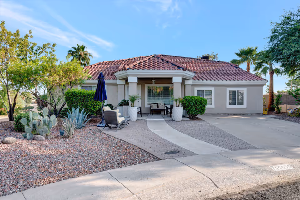 Single-story house with a red tile roof and beige exterior walls. The front yard features desert landscaping with rocks, cacti, and shrubs. There is a paved walkway leading to a covered porch with outdoor seating and a closed blue umbrella. Palm trees and other greenery are visible in the background under a clear blue sky.