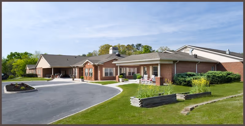 Front exterior of a single-story red-brick assisted living facility with a curved driveway, covered porch and lawn.