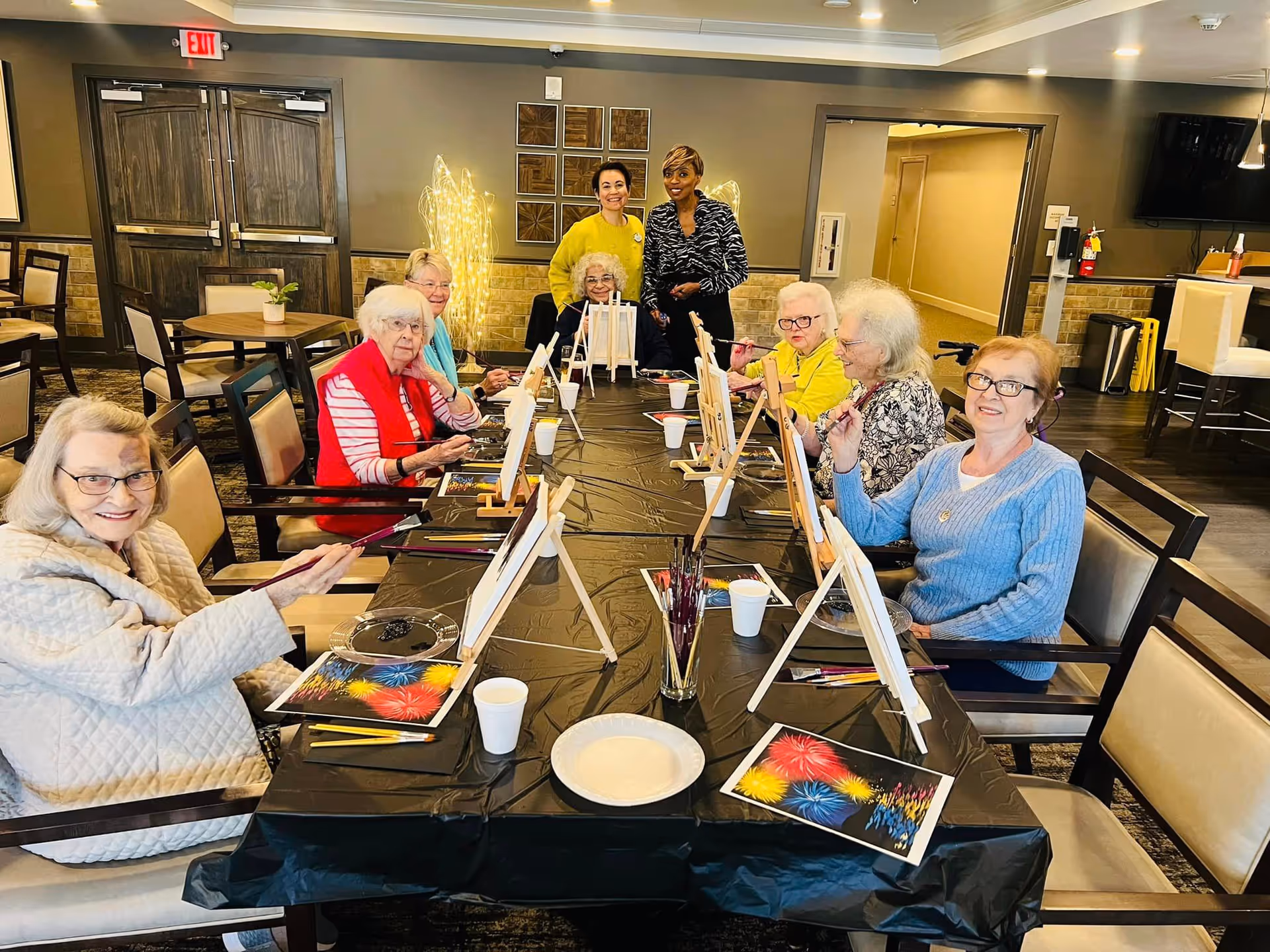 A group of elderly women seated around a long table in a well-lit room, engaged in a painting activity with canvases on easels, paintbrushes, and paint palettes. Two staff members stand behind them, smiling. The room has wooden doors, chairs, and warm lighting.