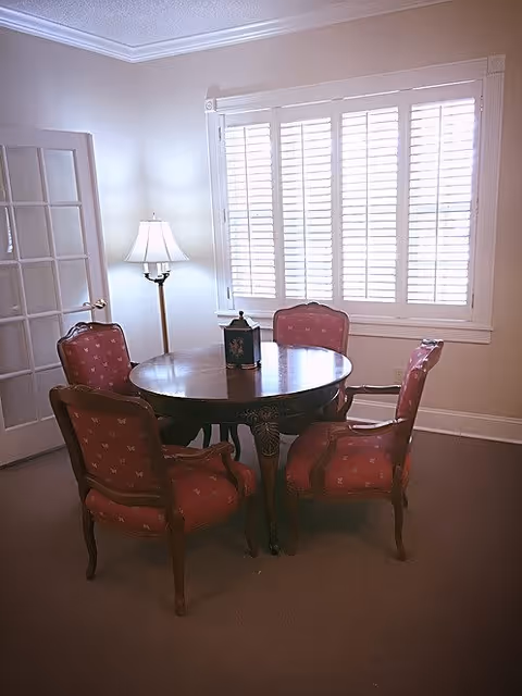 A small round wooden table surrounded by four upholstered chairs with red patterned fabric in a room with beige walls and carpet. A floor lamp with a white shade stands next to a window with white plantation shutters. A glass-paneled door is partially visible on the left side.