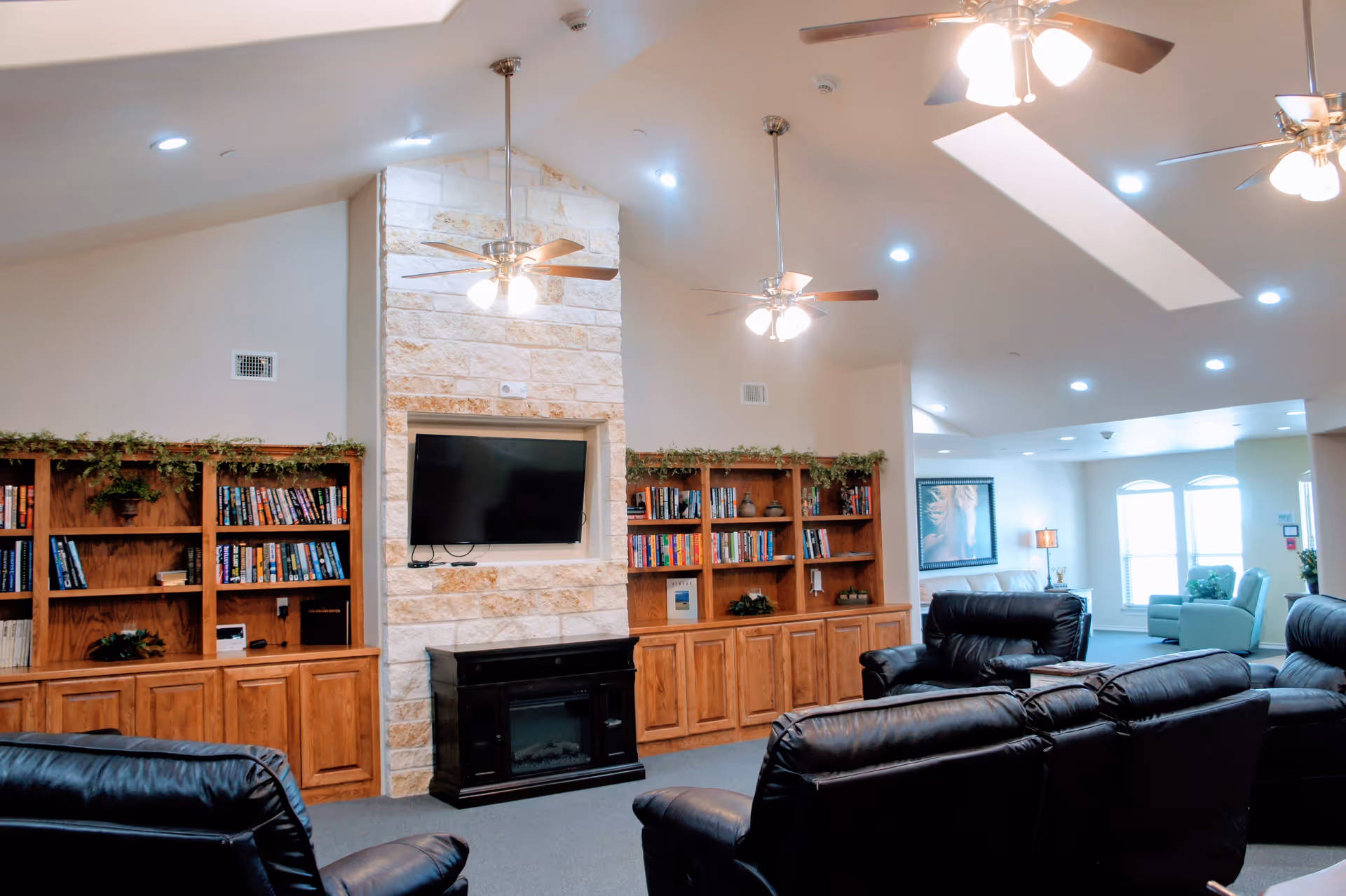 A spacious senior living room with black leather recliners arranged facing a stone fireplace with a mounted flat-screen TV. Wooden bookshelves with books and decorative plants flank the fireplace. The ceiling is vaulted with ceiling fans and recessed lighting. In the background, there is a seating area with light blue chairs near windows letting in natural light.