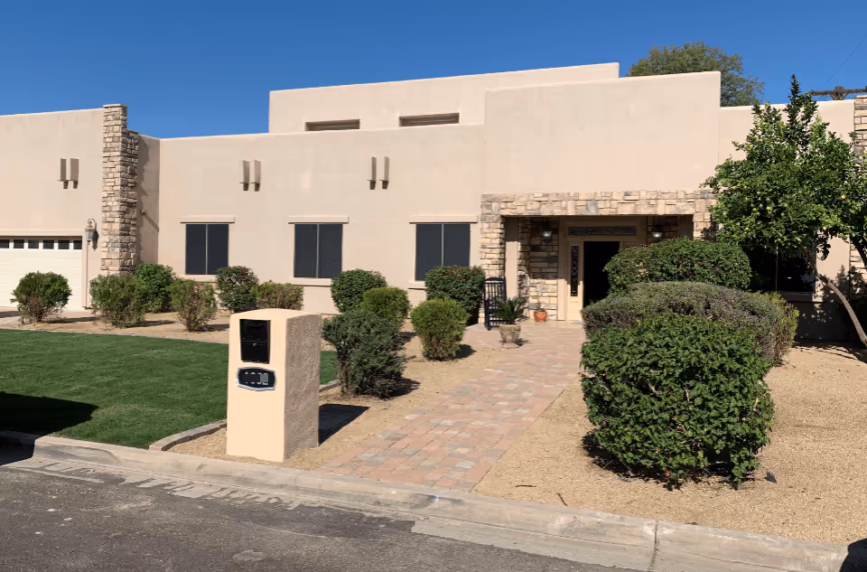 Front entrance of a single-story beige stucco building with stone accents, a paved walkway, mailbox pillar and trimmed bushes.