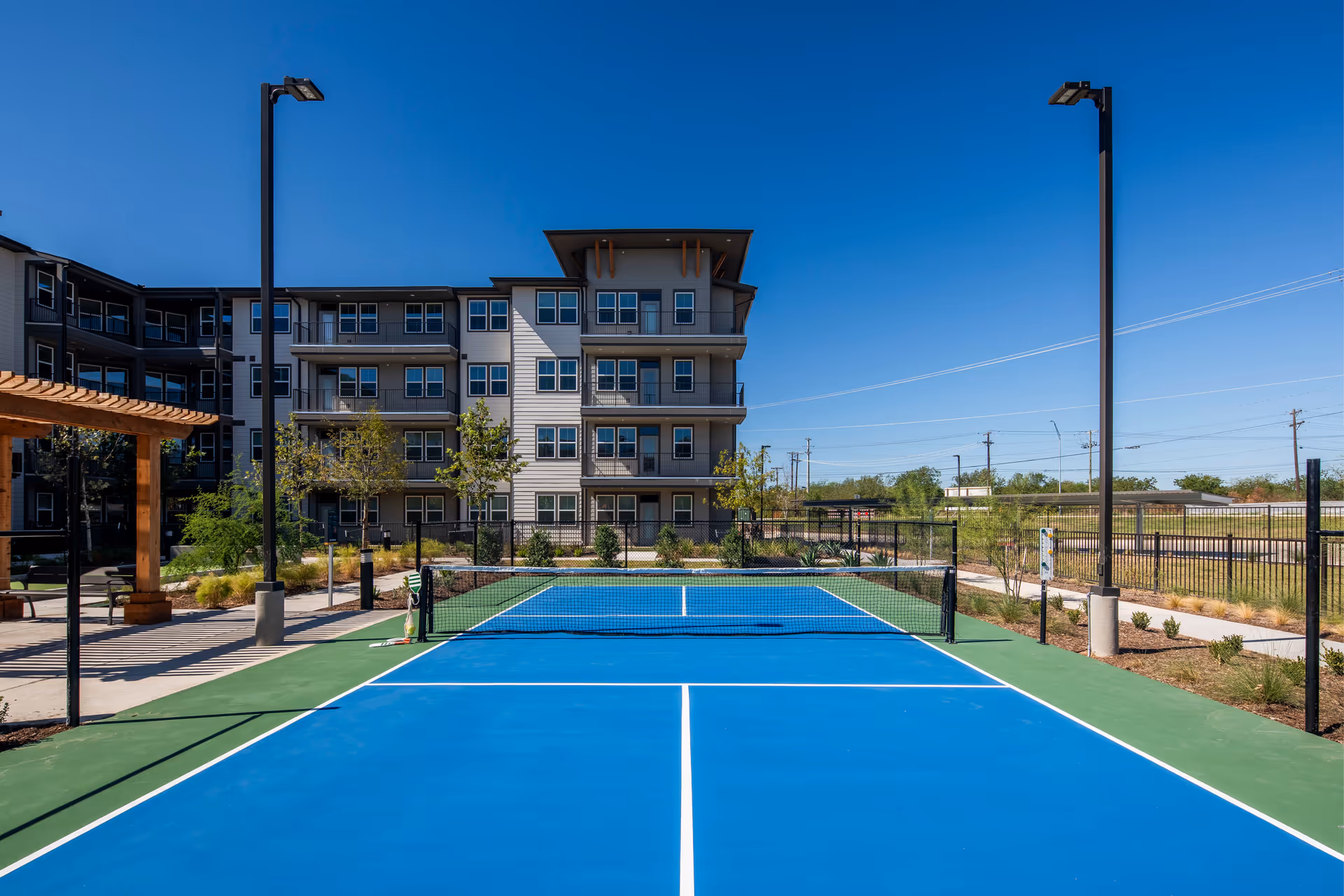A bright blue outdoor pickleball court in front of a four-story senior living building under a clear blue sky.