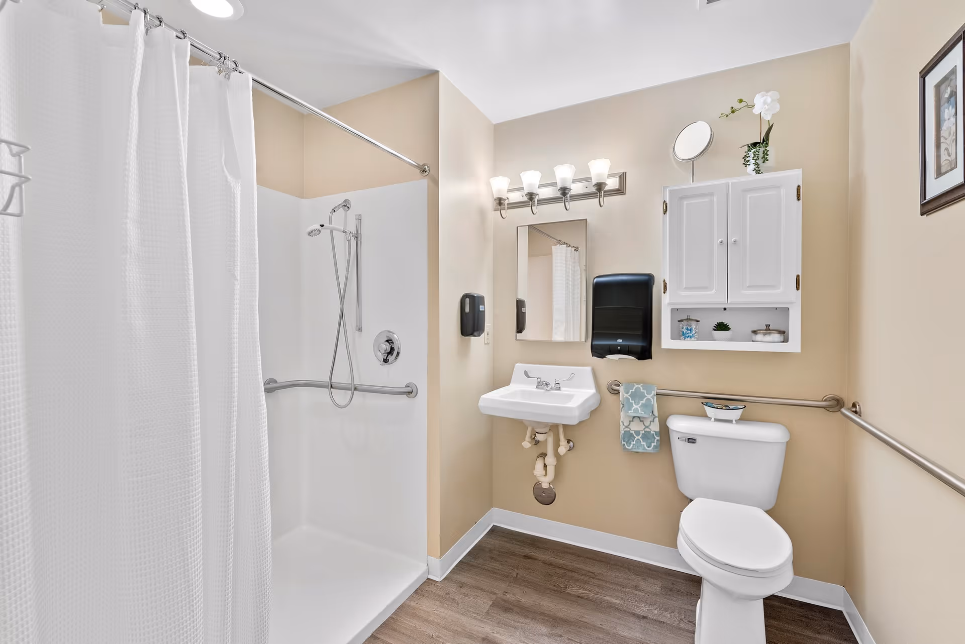 A clean and accessible bathroom with a walk-in shower featuring a white curtain and grab bars, a white sink with a mirror and light fixture above, a white toilet with grab bars on the walls, a white cabinet mounted on the wall with decorative items, and a towel hanging on a rail.