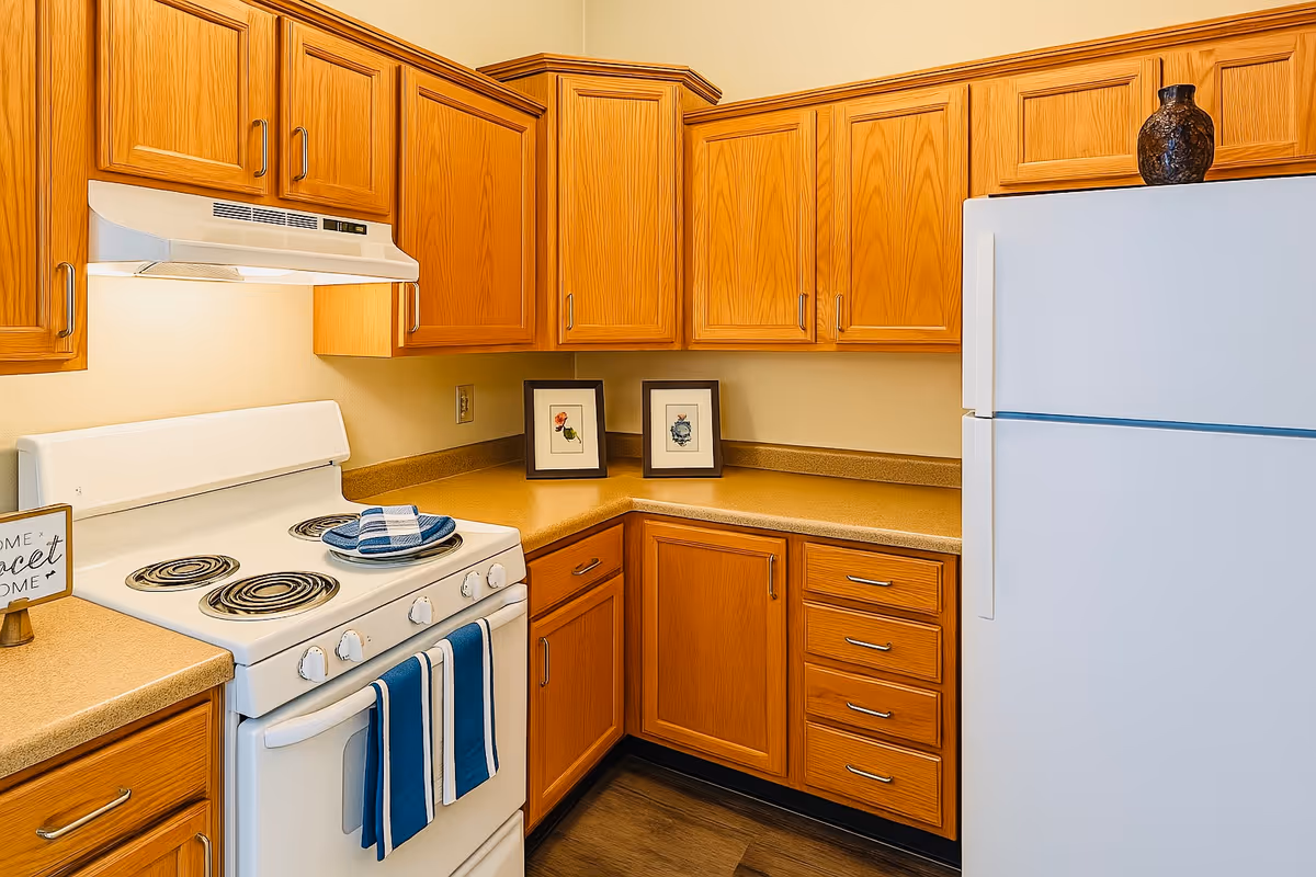 A kitchen with wooden cabinets, a white electric stove with coil burners, a white refrigerator, and a countertop with two framed pictures and a small decorative sign. Blue and white striped towels hang on the stove handle.