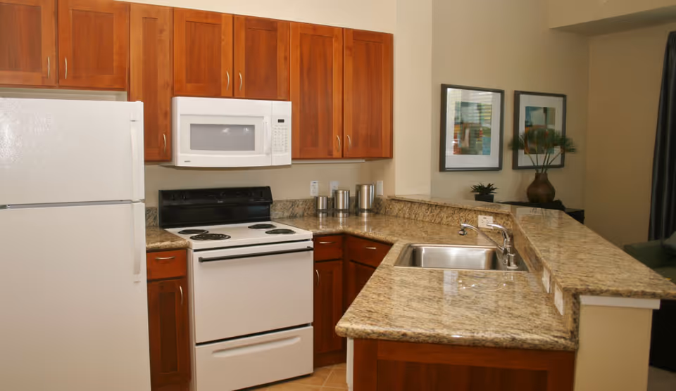 A kitchen with wooden cabinets, a white refrigerator, a white electric stove with an oven, a white microwave above the stove, and a granite countertop with a stainless steel sink. There are two framed pictures and a decorative vase with greenery on a counter in the background.