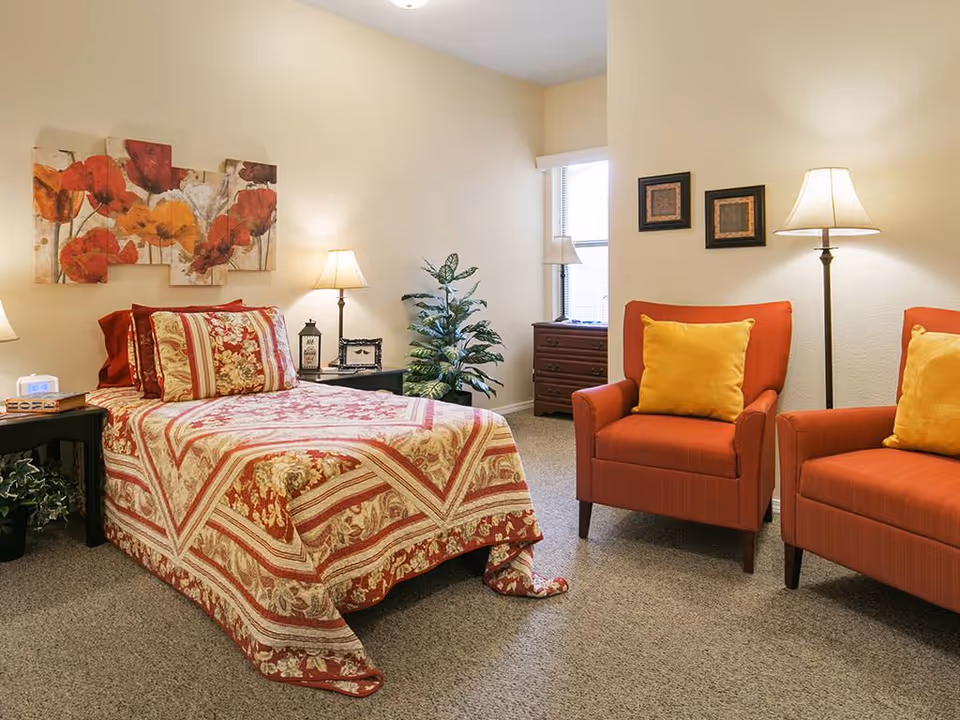A warmly decorated bedroom with a single bed covered in a red floral quilt, two orange armchairs with yellow pillows, lamps, and wall art.