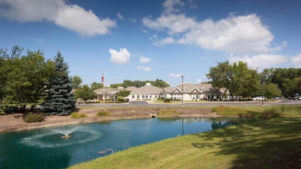 One-story senior living building seen across a pond with a small fountain, trees, an American flag, and a blue sky with clouds.