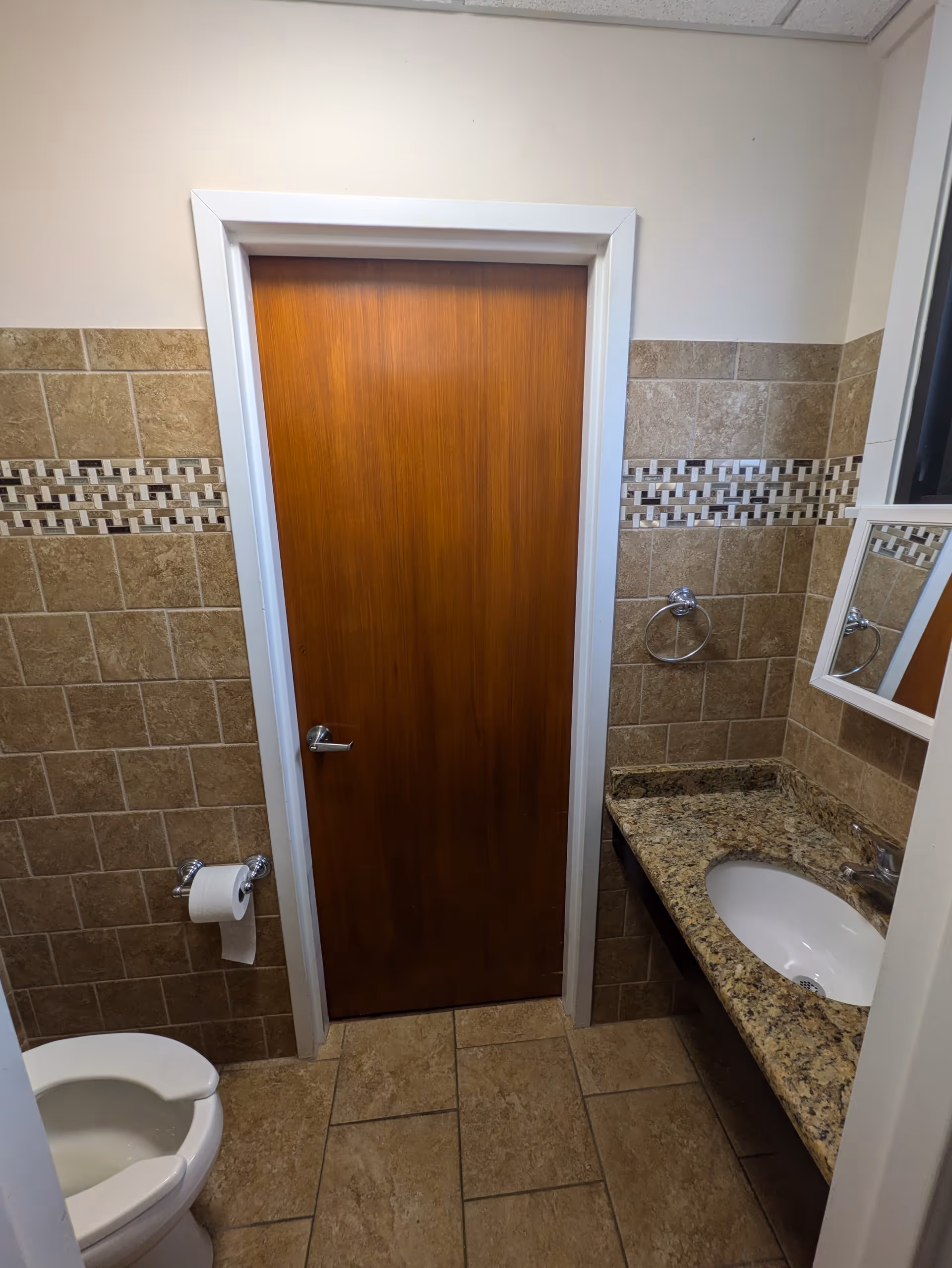 Small bathroom with beige tiled walls and floor, a wooden door in the center, a white toilet with a toilet paper holder on the left, and a granite countertop with a sink and a mirror on the right.