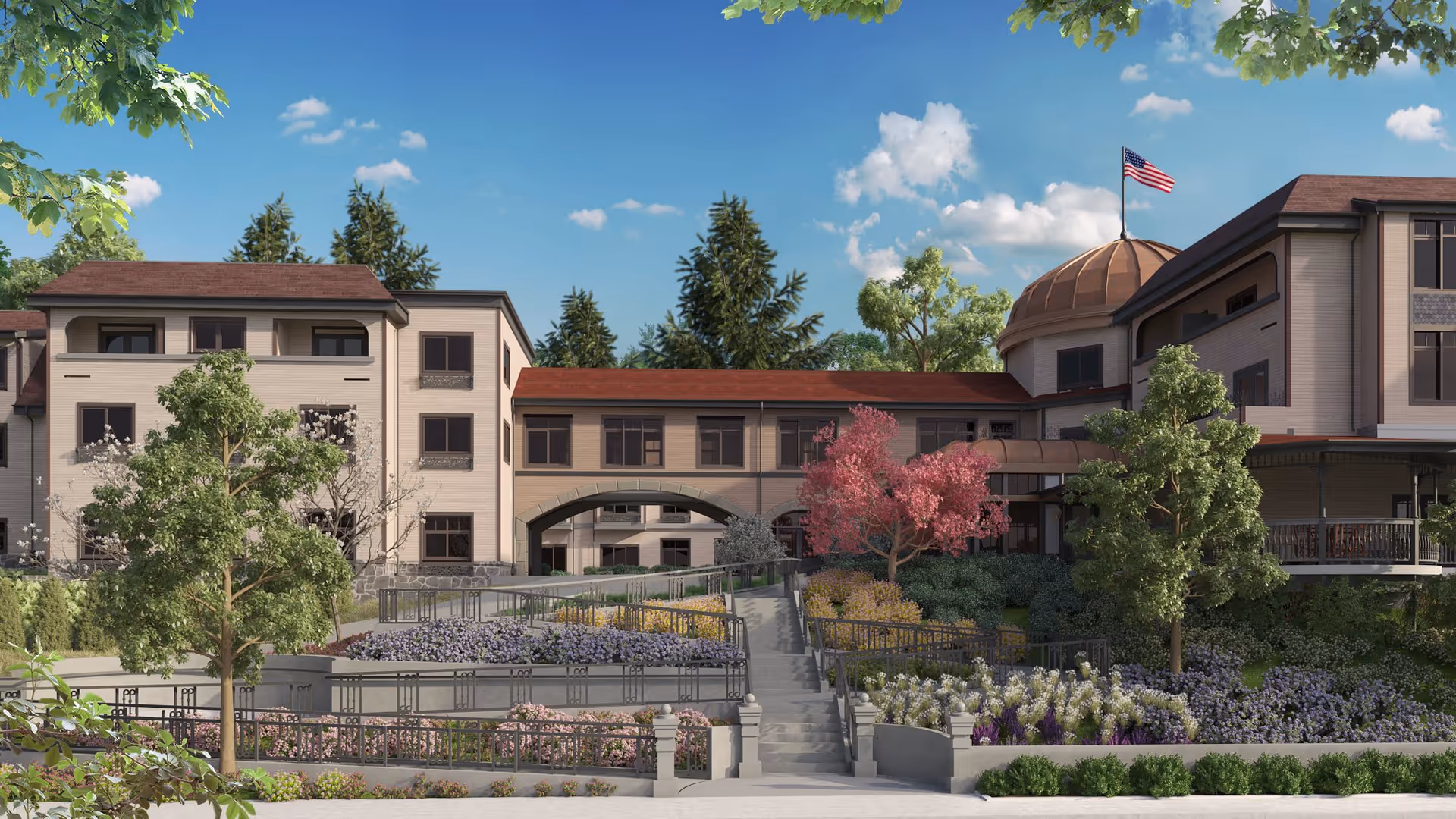 Exterior view of a senior living facility building with beige walls and red roofs, surrounded by landscaped gardens with various trees and flowering plants. A staircase and ramp lead up to the entrance, and an American flag flies atop a dome structure on the right side of the building under a blue sky with some clouds.