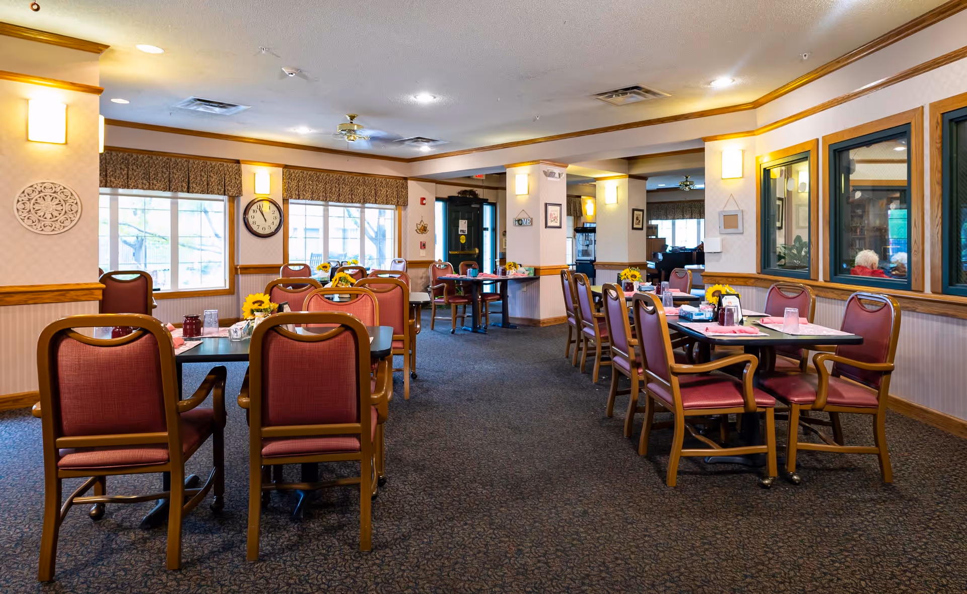 A spacious dining room in an assisted living facility with multiple tables and chairs arranged neatly. The tables have placemats, glasses, and small flower arrangements. The room has large windows with valances, wall sconces for lighting, and a clock on the wall. The carpet is patterned, and the walls have wood trim and light-colored wallpaper.