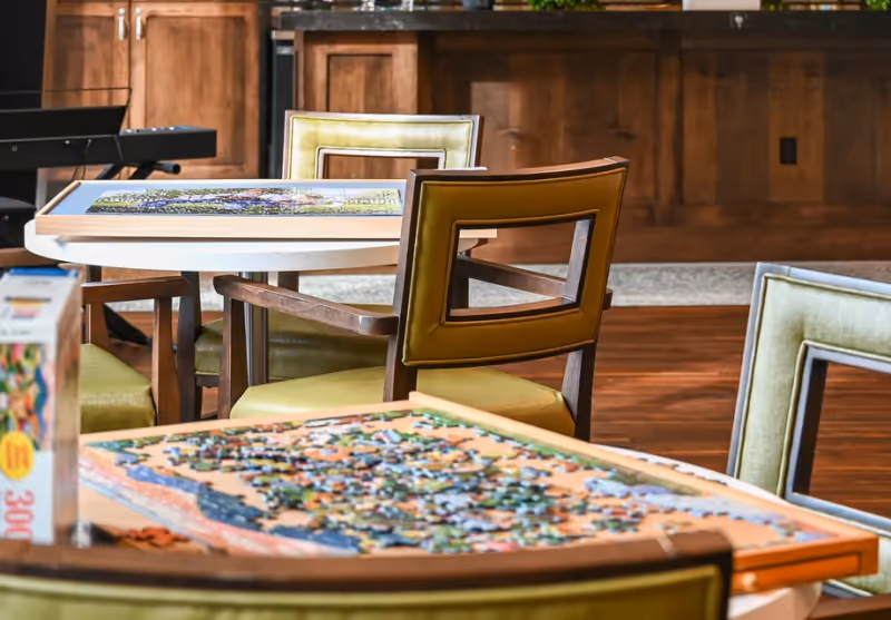 Tables with partially assembled jigsaw puzzles and green-upholstered wooden chairs in a communal activity room.