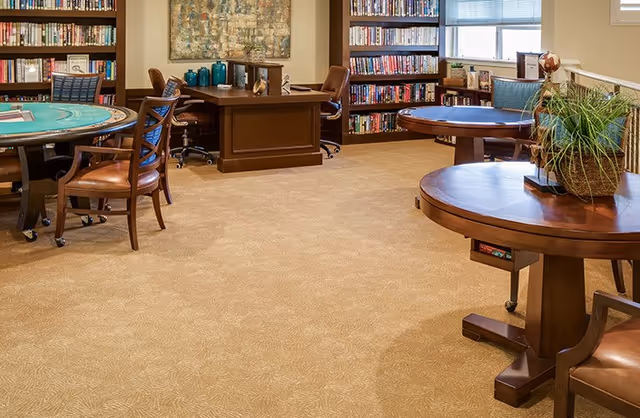 A cozy room with carpeted floor featuring multiple wooden tables and chairs, bookshelves filled with books along the walls, a desk with a chair, and a large window letting in natural light. There is a plant on one of the tables and decorative items on the desk and shelves.