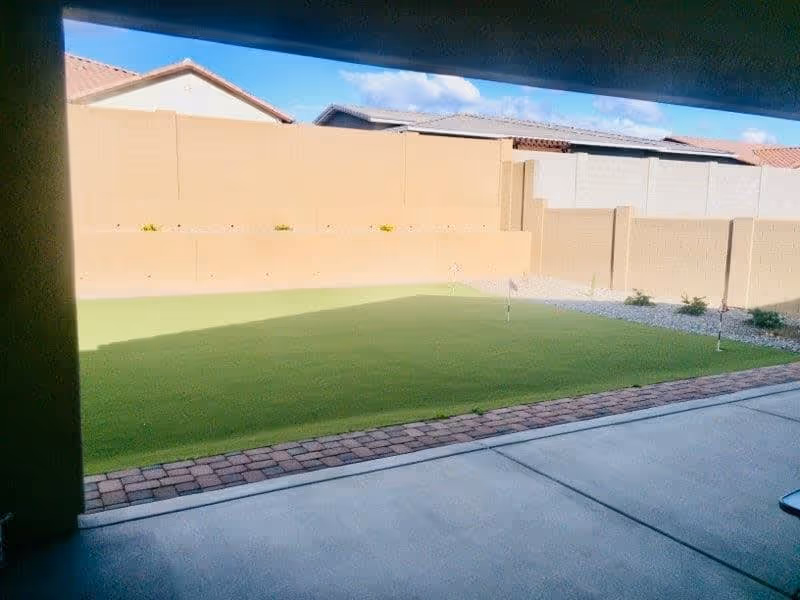 View of a backyard area with a putting green, small flags, and a brick border, enclosed by beige and gray block walls with rooftops visible in the background under a blue sky with some clouds.