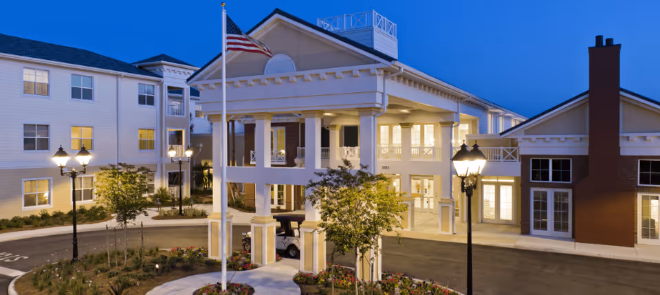 Exterior front entrance of a senior living facility at dusk with a covered porte-cochère, American flag, lampposts, and illuminated windows.