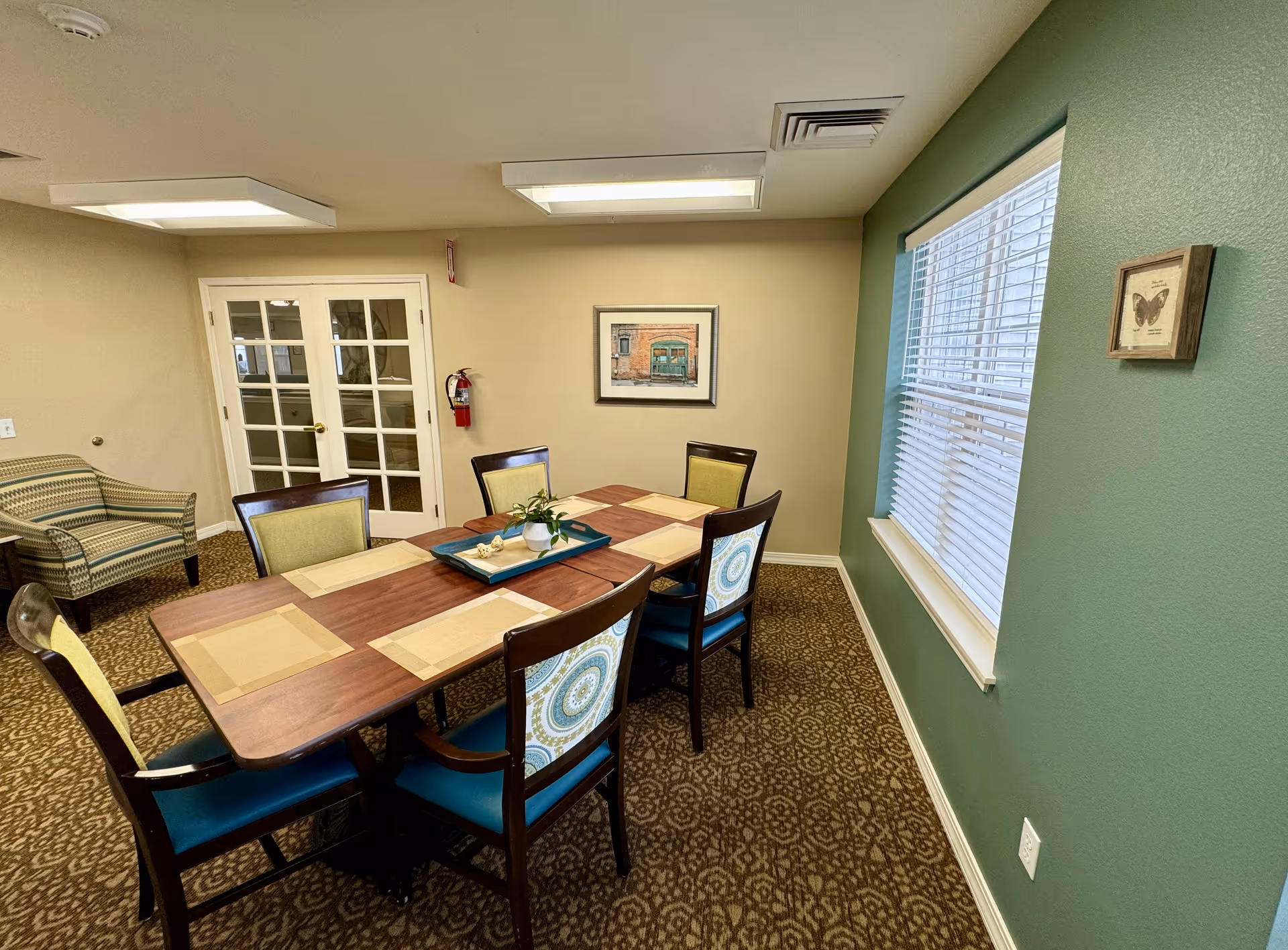 A dining room in a senior living facility with a wooden table set for six, featuring placemats and a decorative centerpiece. The room has patterned carpet, a green accent wall with a window, beige walls, a framed picture, a striped armchair, and double glass doors.