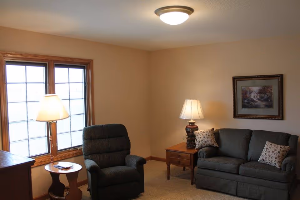 A cozy living room with a dark upholstered armchair and a matching sofa with floral pillows. There are two wooden side tables, each with a lamp, one near the window and one beside the sofa. A framed painting hangs on the wall above the sofa. The room has beige walls and carpeted floor.