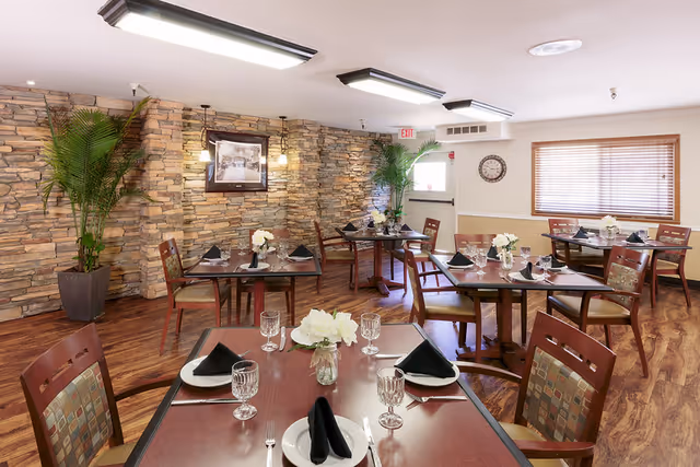 A dining room with several wooden tables set with white plates, black folded napkins, clear water glasses, and silverware. Each table has a small vase with white flowers. The room features a stone accent wall with framed artwork, two large potted plants, wooden flooring, and a window with wooden blinds. Ceiling lights illuminate the space.