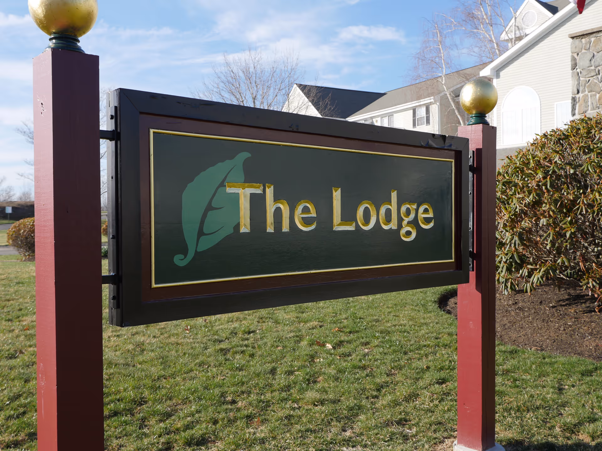 A wooden sign with gold lettering that reads 'The Lodge' mounted on two red posts with gold spherical finials, set on a grassy area with a building and bushes in the background under a blue sky.