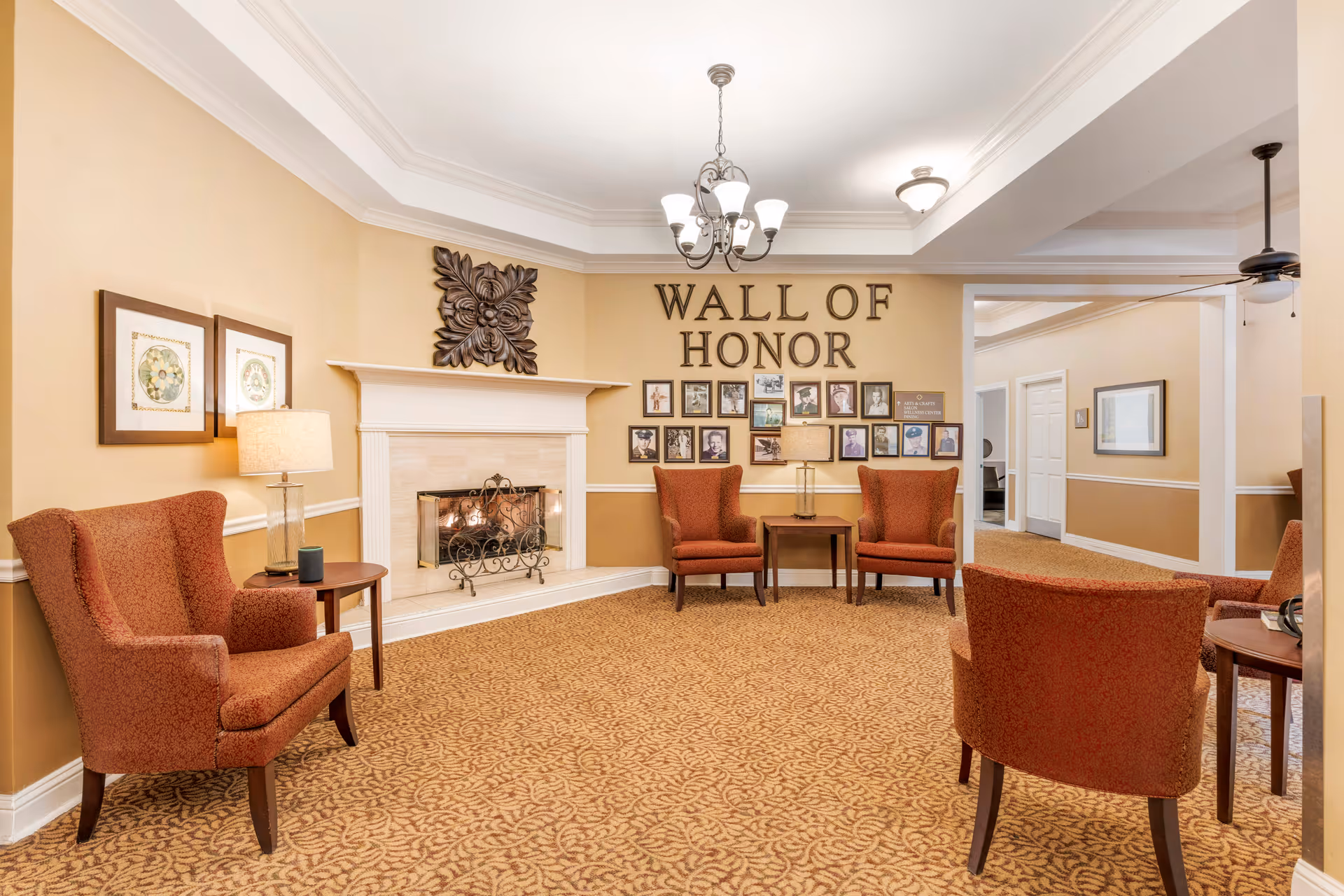 A cozy sitting area with four orange upholstered armchairs arranged around two small wooden tables. There is a fireplace with a decorative metal screen and a large floral wall decoration above it. On the wall behind two of the chairs is a 'Wall of Honor' display featuring framed photographs. The room has beige walls, patterned carpet, and ceiling light fixtures.
