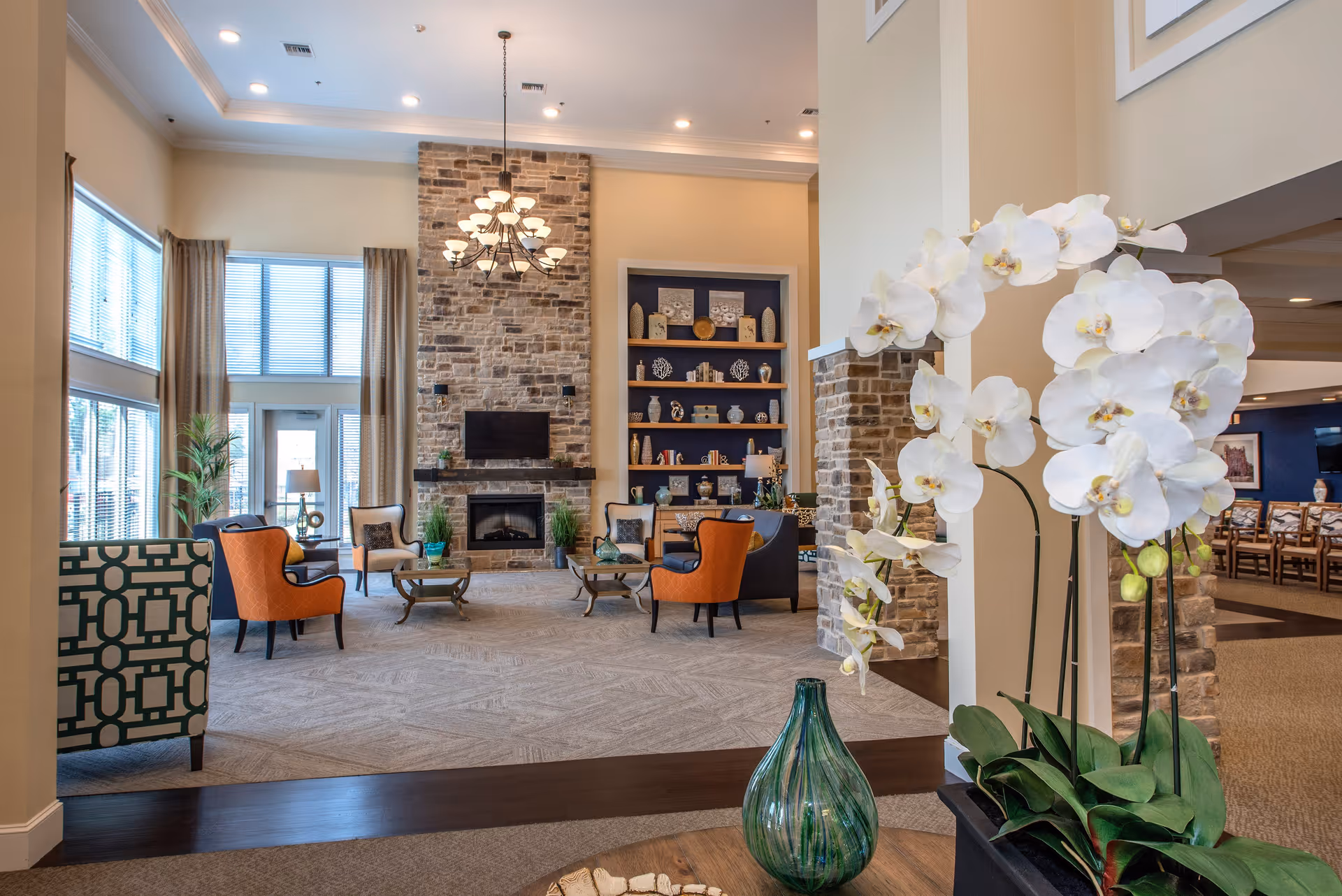 A spacious and well-lit living room area with a stone fireplace and a mounted TV. The room features comfortable seating including orange and patterned armchairs arranged around small tables. Large windows with beige curtains allow natural light to fill the space. Decorative shelves with various ornaments are visible on the wall next to the fireplace. In the foreground, there is a white orchid plant and a green glass vase on a wooden table.
