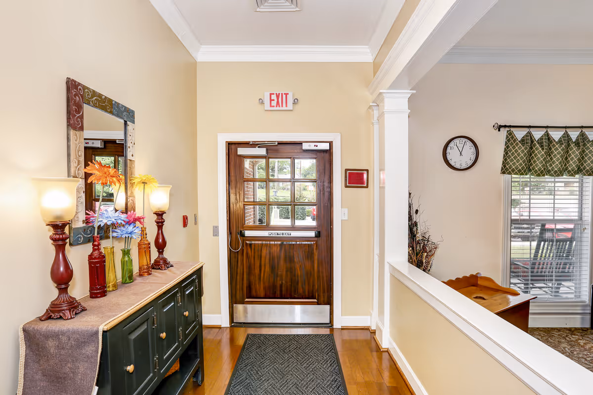 Interior view of an entryway in a senior living facility with a wooden door featuring glass panels and an exit sign above it. To the left, there is a dark green cabinet with a brown runner, decorated with colorful vases holding artificial flowers and two lamps with frosted glass shades. A large decorative mirror hangs above the cabinet. To the right, there is a partial view of a room with a window covered by a green valance, a wall clock, and a wooden rocking chair visible outside the window.