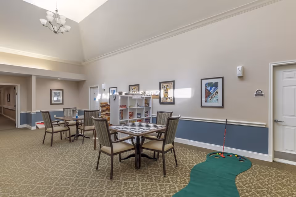 A well-lit common area in a senior living facility featuring two tables with chairs, one table with a checkerboard pattern, a shelf with board games and puzzles, framed artwork on the walls, a mini putting green with golf balls and a putter, and a hallway leading to other rooms.