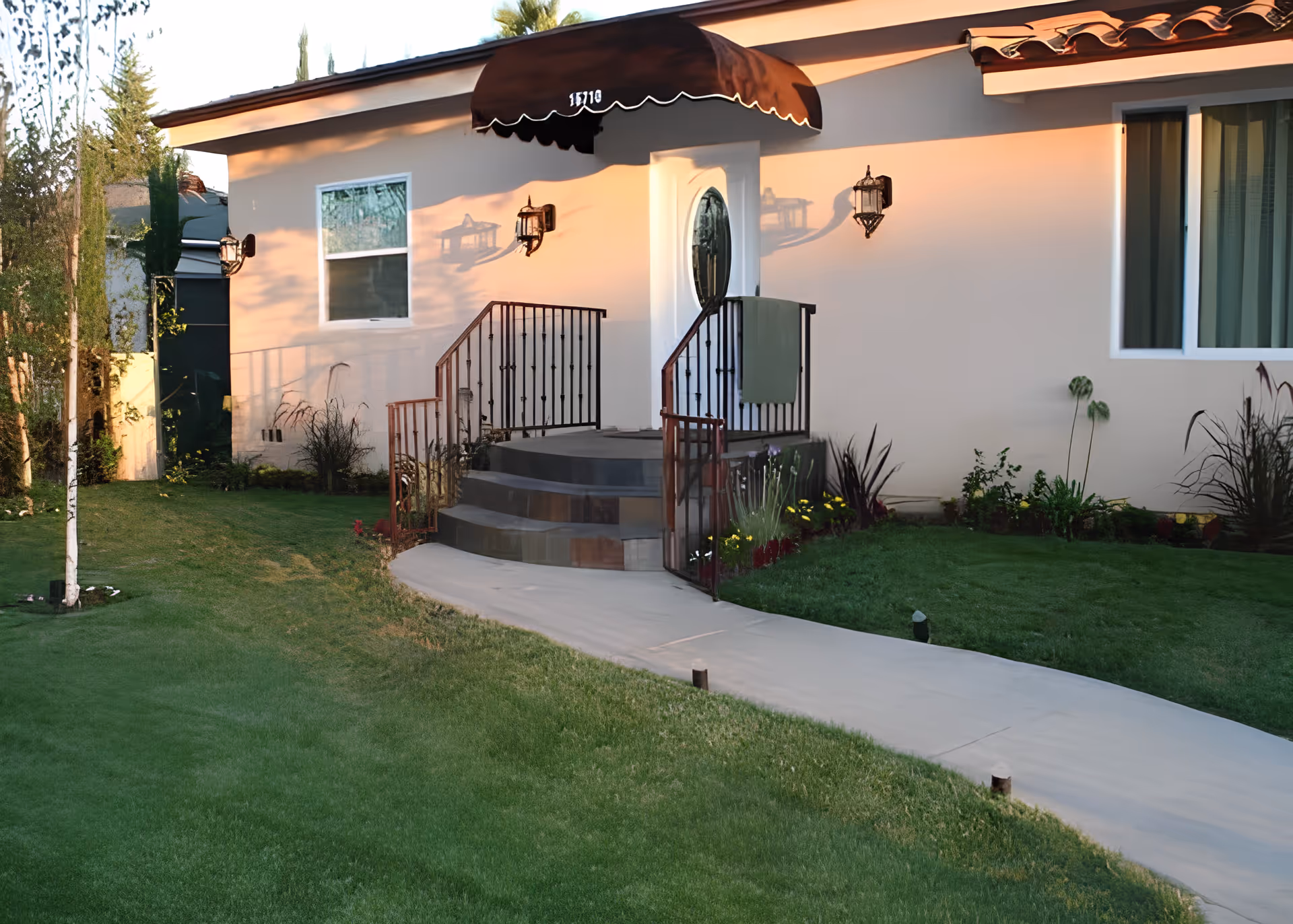 Front entrance of a single-story building with a small staircase leading to a white door under a brown awning. The building is surrounded by a well-maintained lawn and garden with various plants and flowers. There are two wall-mounted lantern-style lights on either side of the door.