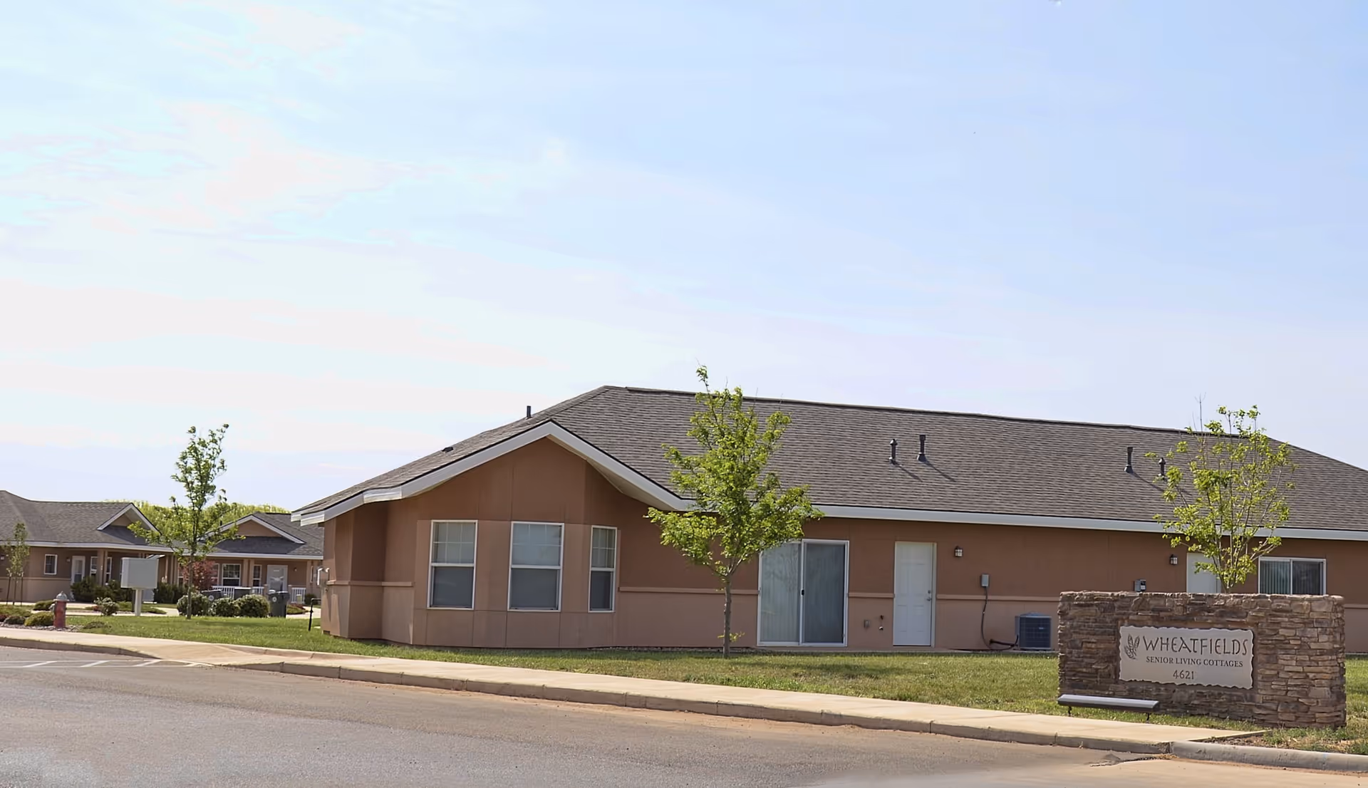 Exterior view of a single-story senior living facility building with a brown roof and beige walls, surrounded by small trees and a well-maintained lawn. A stone sign in front reads 'Wheatfields Senior Living Cottages 4621'.