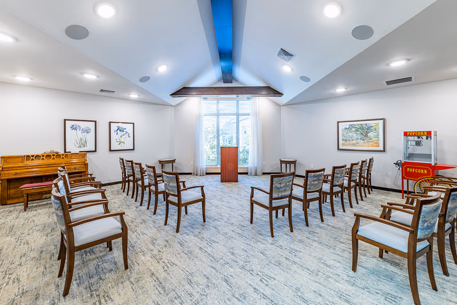 A bright, spacious room with a high vaulted ceiling featuring a wooden beam. The room is arranged with wooden chairs in a semi-circle facing a wooden podium in front of a large window with sheer curtains. On the left side, there is an upright piano with a bench, and on the right side, a popcorn machine and framed artwork hang on the walls. The carpet has a light, textured pattern.