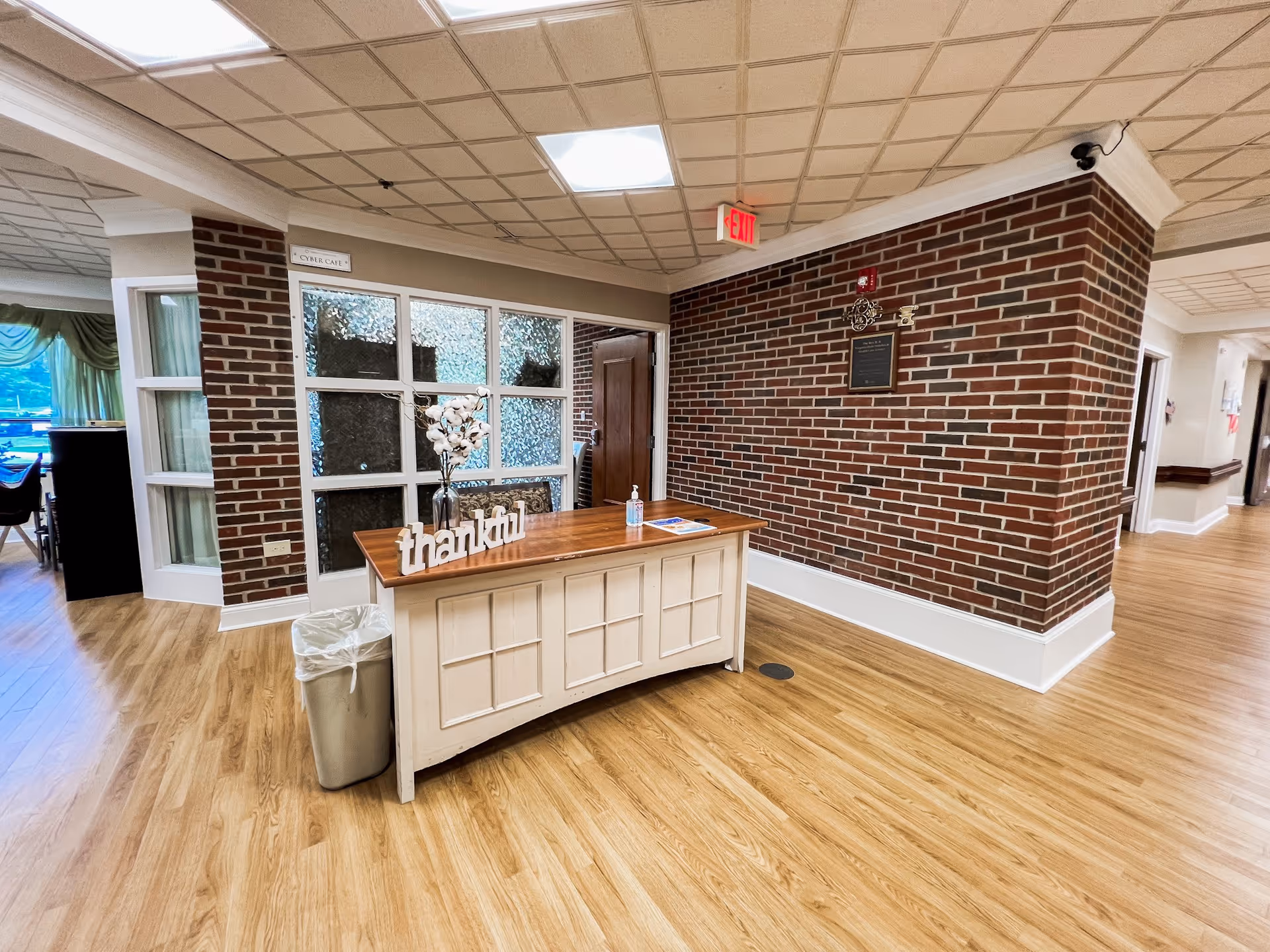 Interior view of a senior living facility hallway with wood flooring and brick walls. A white reception desk with a wooden top is positioned near a corner, decorated with a vase of flowers and a 'thankful' sign. There is a trash can beside the desk and a hand sanitizer bottle on top. To the left, a room with large windows and green curtains is partially visible. The ceiling has recessed lighting and an exit sign is mounted above a door in the brick wall.