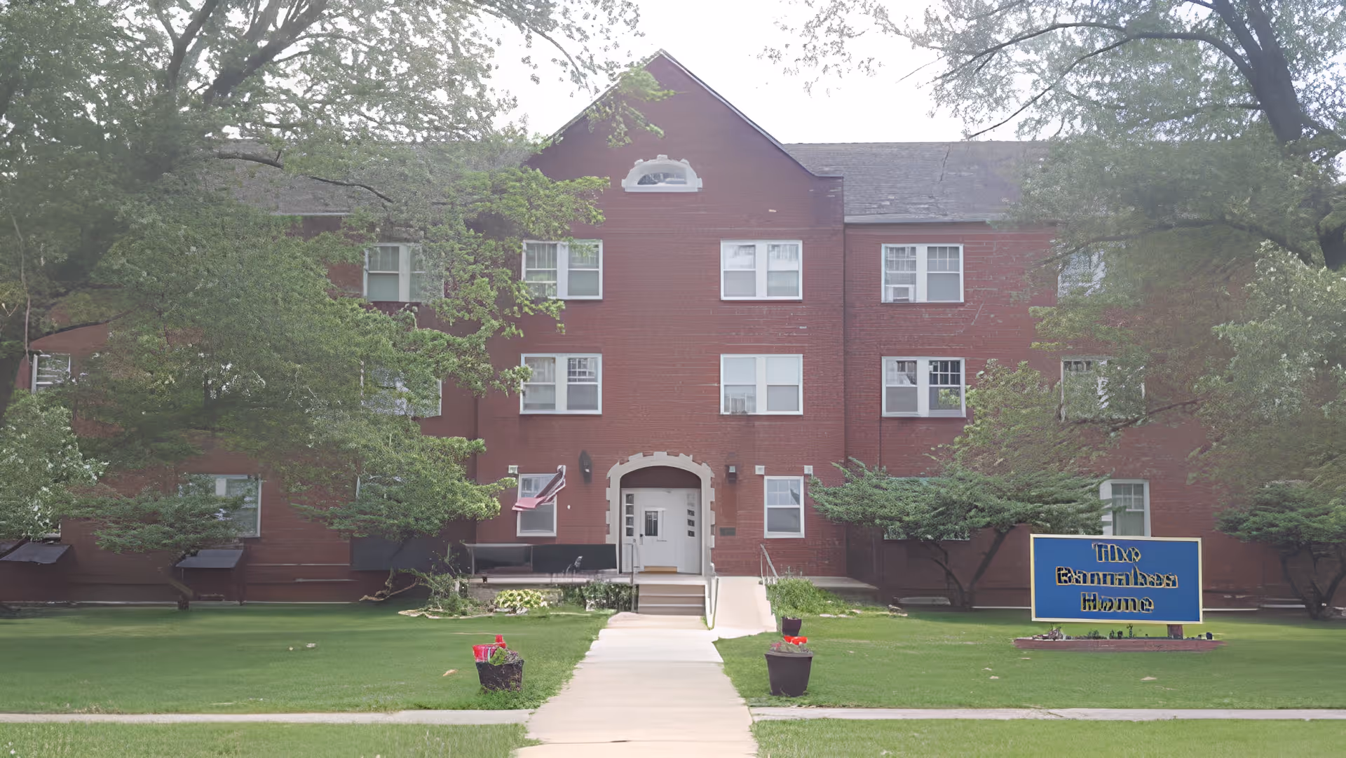 Front view of a three-story red-brick senior living building with a central entrance, walkway, lawn, and a sign.