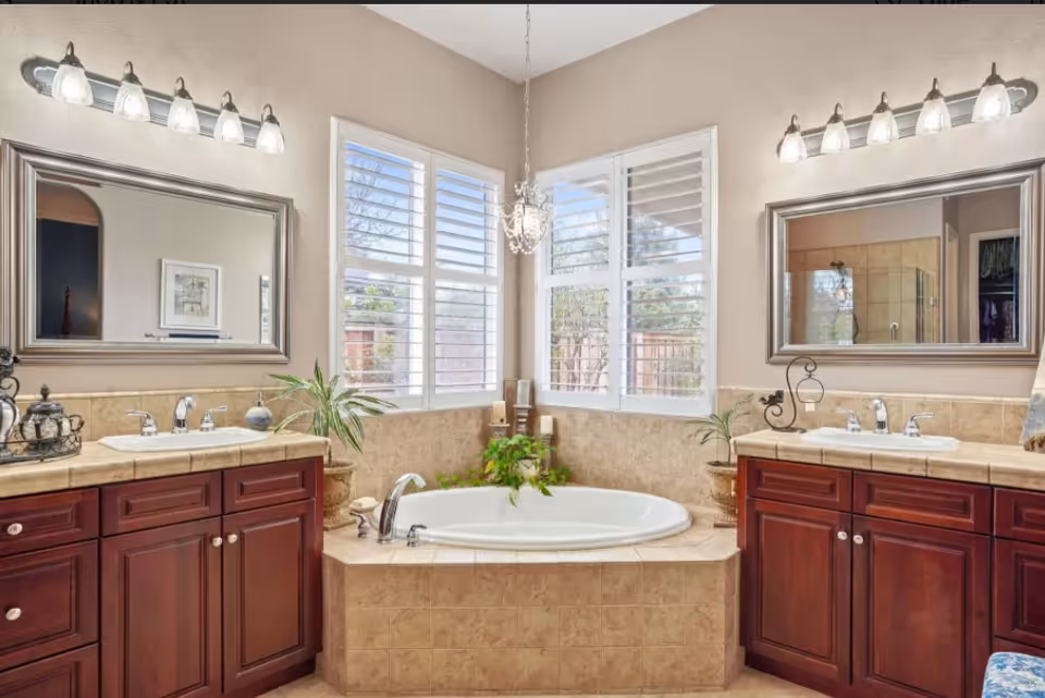 A spacious bathroom featuring a large corner bathtub surrounded by beige tiles, flanked by two wooden vanities with sinks and mirrors above each. The room has two large windows with white shutters allowing natural light to enter. There are decorative plants and candles around the bathtub, and light fixtures with multiple bulbs above each mirror.