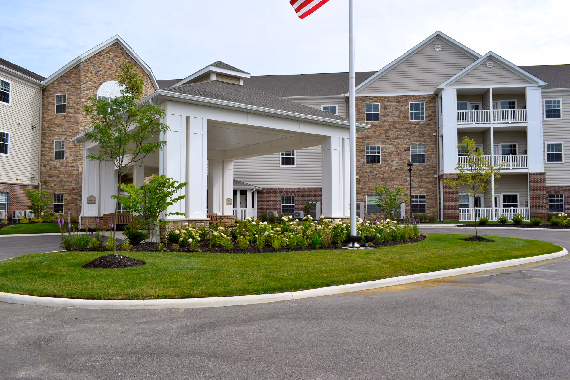 Senior living facility with stone facade, white porte-cochere, flagpole, and manicured landscaping.