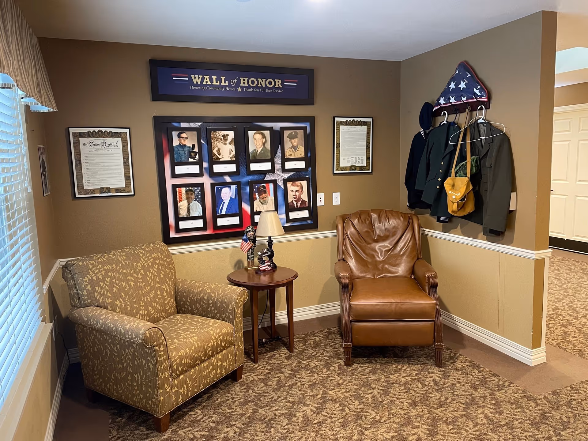 A cozy corner in a senior living facility with two armchairs, one patterned fabric and one brown leather, separated by a small round wooden table with patriotic decorations. On the wall behind the chairs is a 'Wall of Honor' display featuring photos and names of community heroes, along with framed documents and a rack holding military uniforms and a folded American flag.