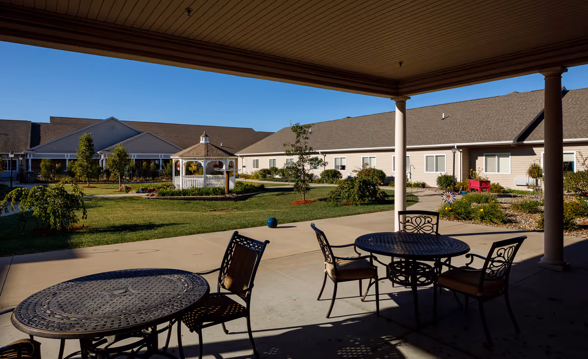 Covered patio with metal tables and chairs overlooking a landscaped courtyard with a white gazebo and single-story assisted living buildings.